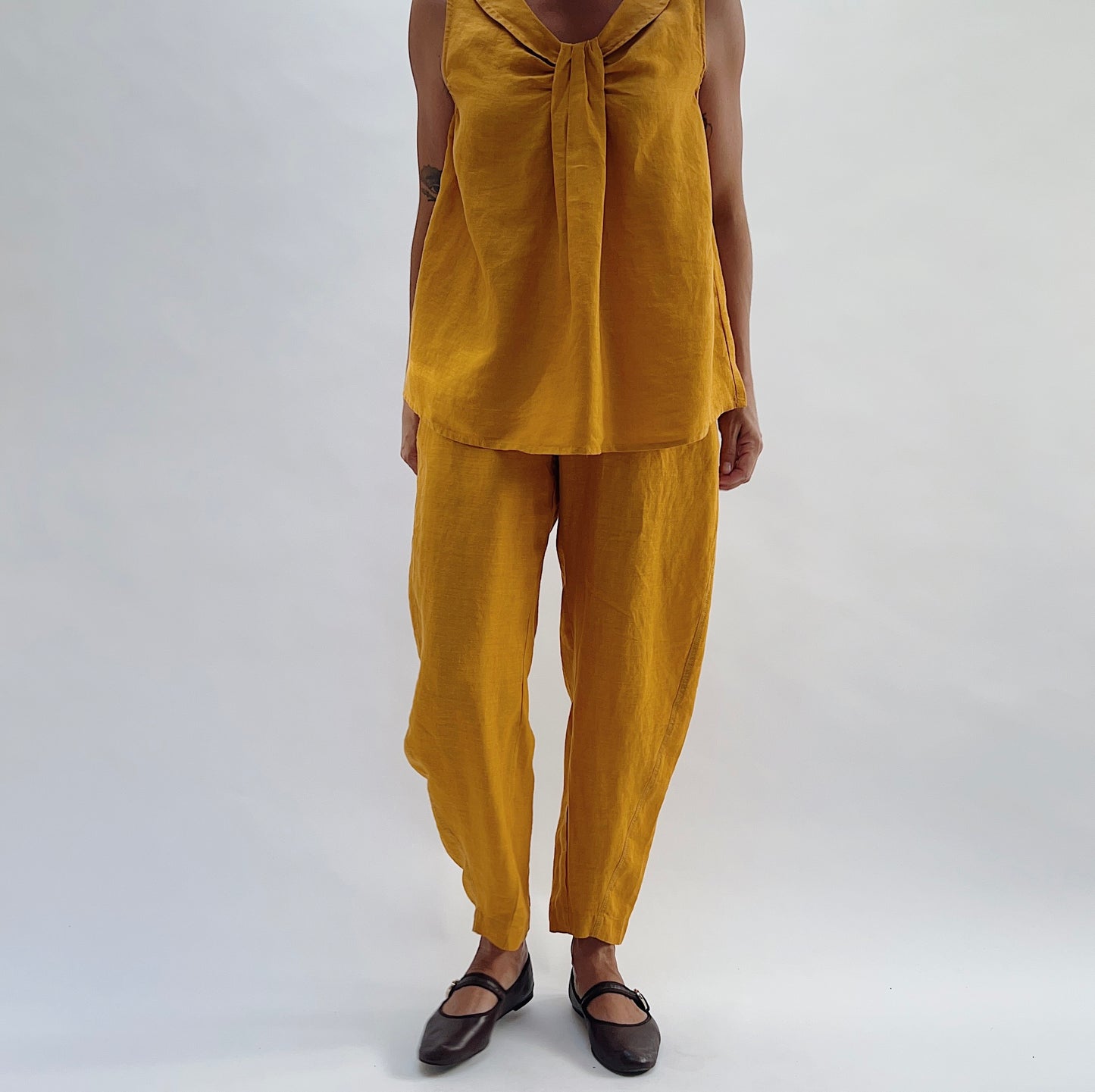 Full-length front view of the Seamed Taper Pant in Tumeric by Yuvita, showing the relaxed tapered silhouette in golden yellow linen, styled with a matching tie-front linen top and dark Mary Jane flats