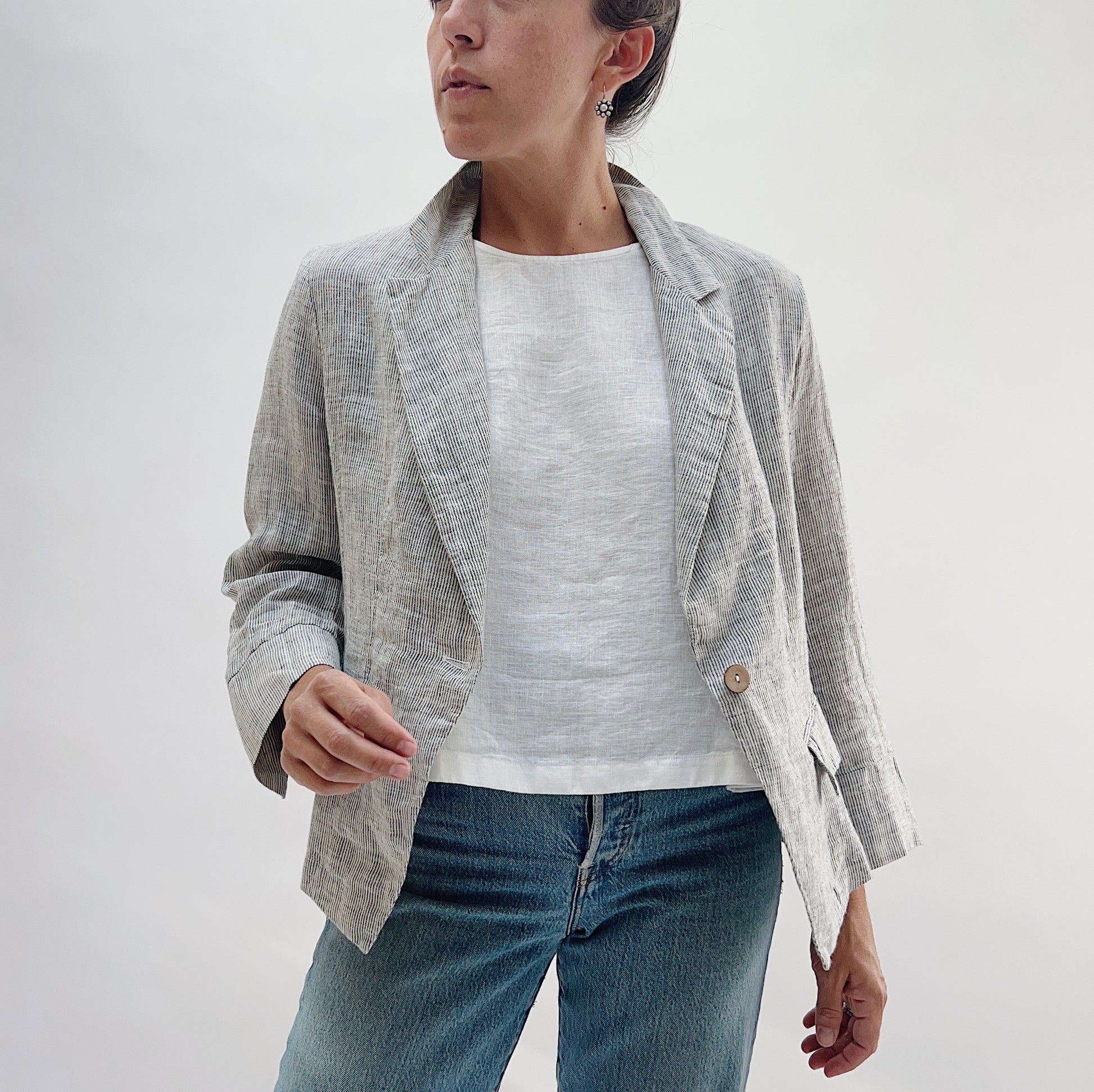 Front three-quarter view of the Linen Blazer in Grey Pinstripe by Yuvita, showing the open lapel, single-button closure, and flap pockets, styled with a white linen tee and blue jeans
