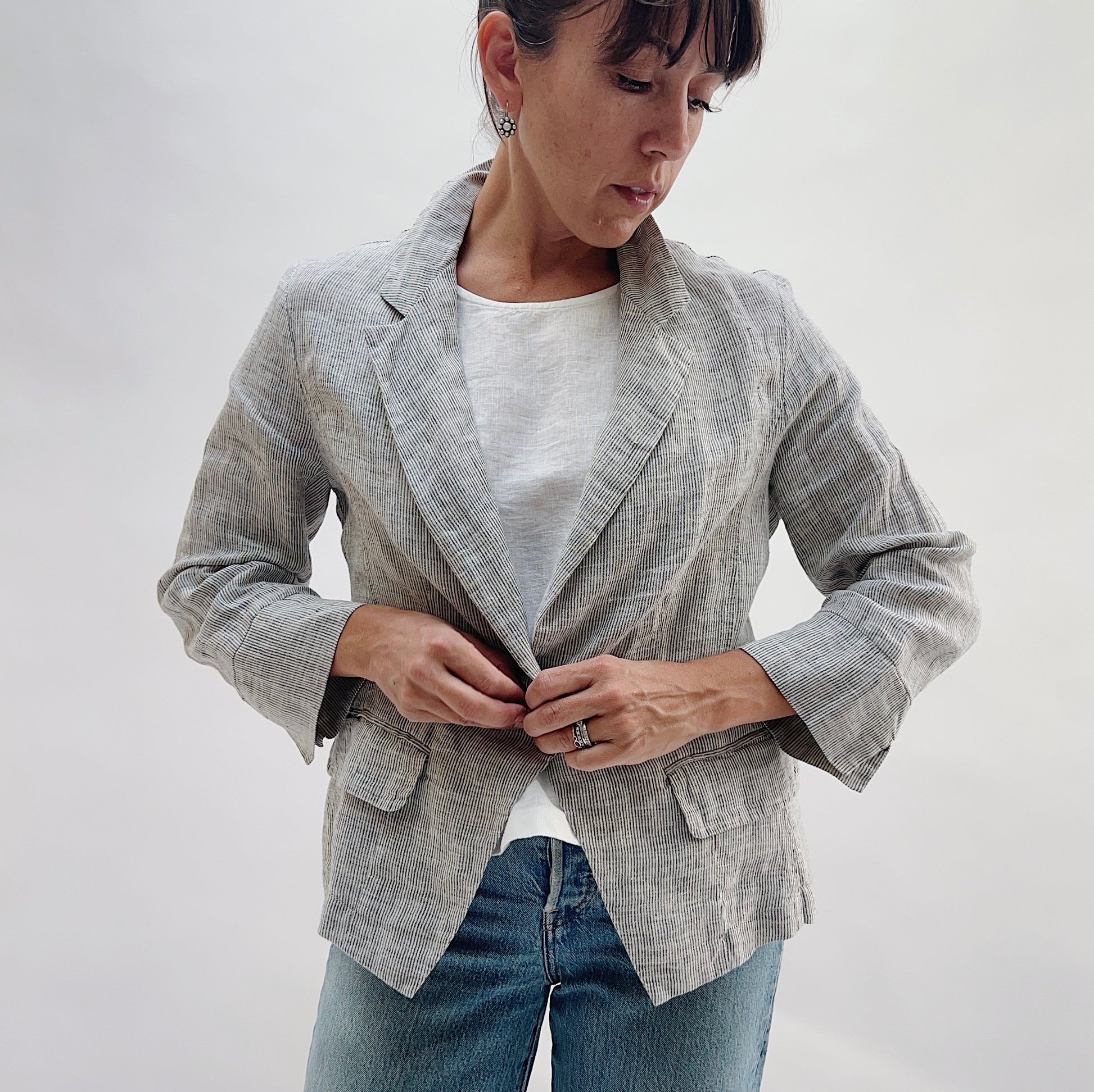 Front view of the Linen Blazer in Grey Pinstripe by Yuvita being buttoned, showing the single-button closure, notched lapel, and flap pockets, styled with a white linen tee and blue jeans
