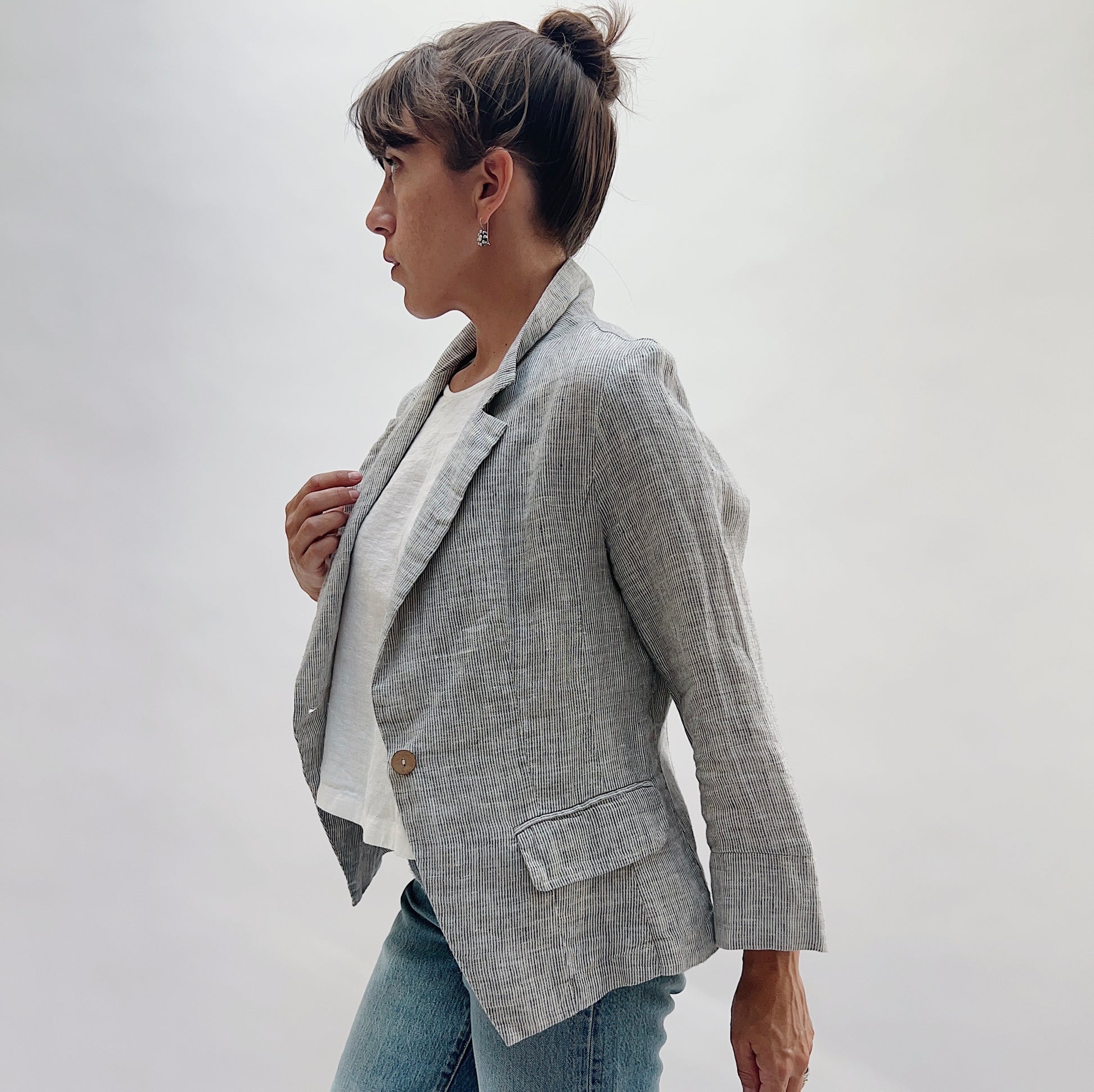 A woman wearing a light gray linen-textured blazer over a white t-shirt and blue jeans, shown from a side profile against a plain light gray studio background.