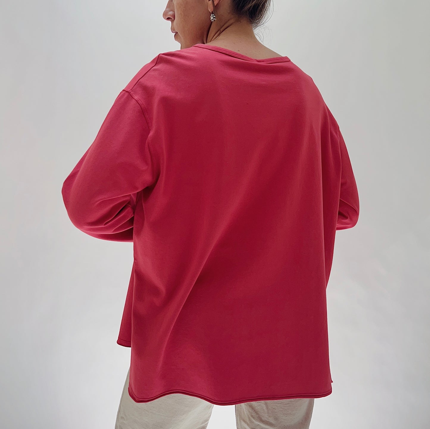 North Star Base Cotton Jersey Beach Tee in Cayenne — rear view showing the wide boxy back, dropped shoulders, long sleeves, and curved hem with side slits in cayenne red, worn with cream trousers on a white background