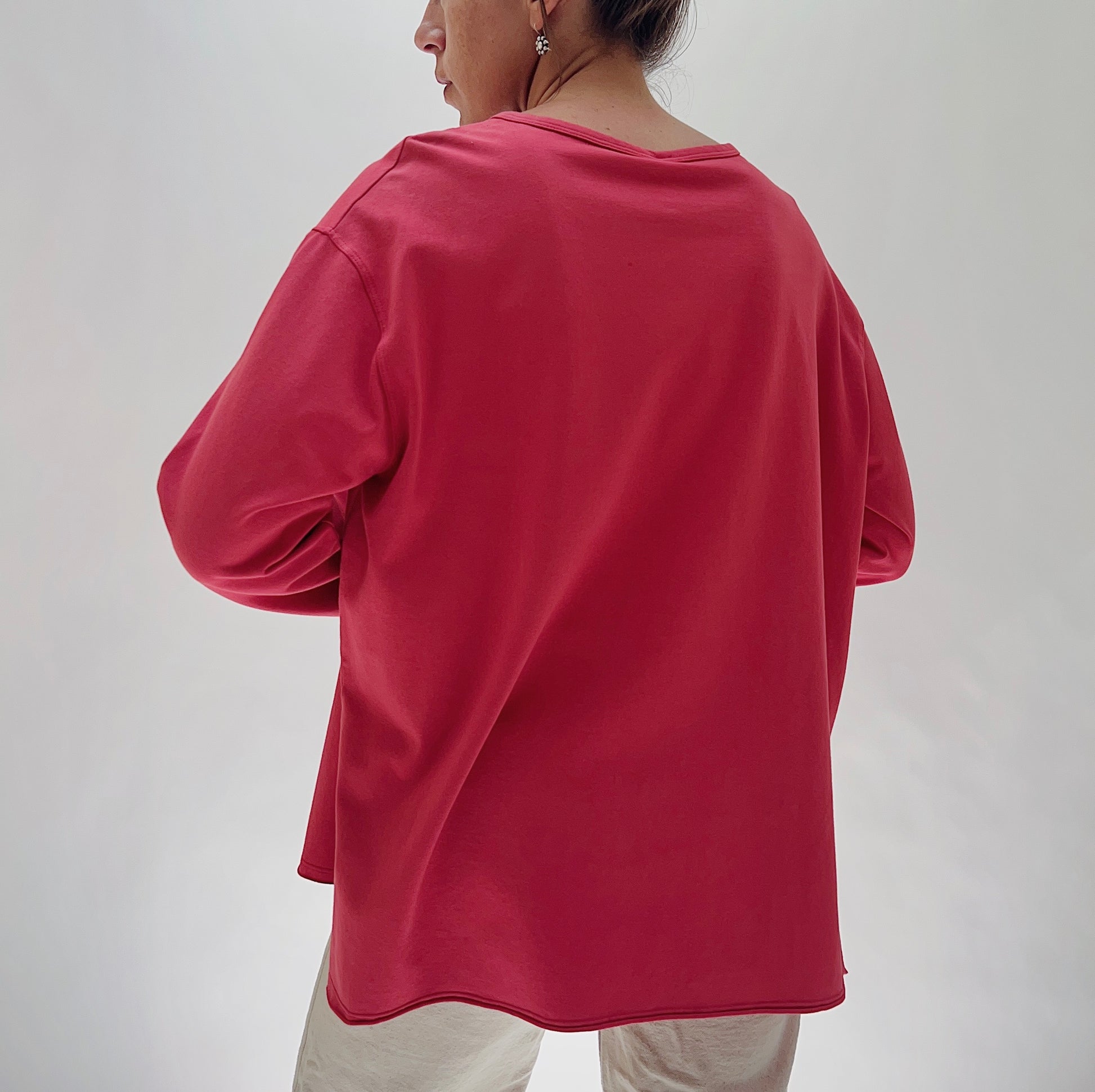 North Star Base Cotton Jersey Beach Tee in Cayenne — rear view showing the wide boxy back, dropped shoulders, long sleeves, and curved hem with side slits in cayenne red, worn with cream trousers on a white background