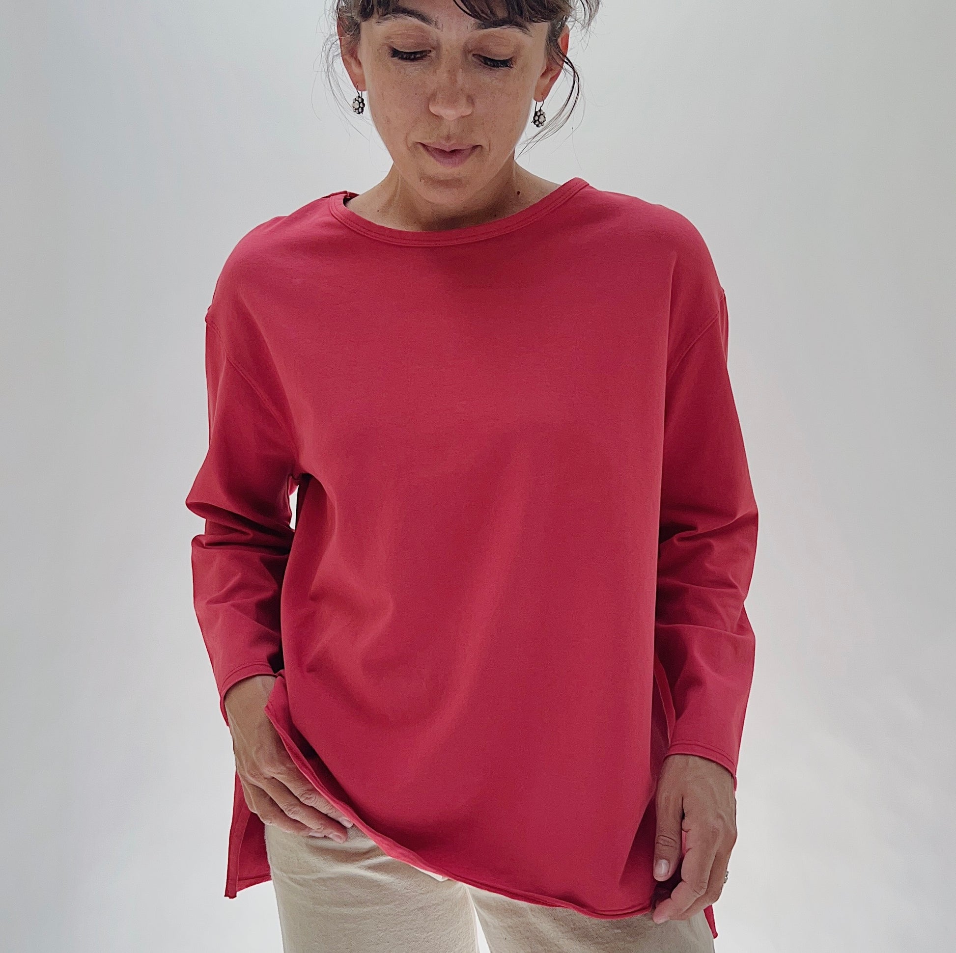 North Star Base Cotton Jersey Beach Tee in Cayenne — front three-quarter view with hands pulling at the hem, showing the relaxed oversized silhouette, crew neckline, and side slits in cayenne red, worn with cream trousers on a white background
