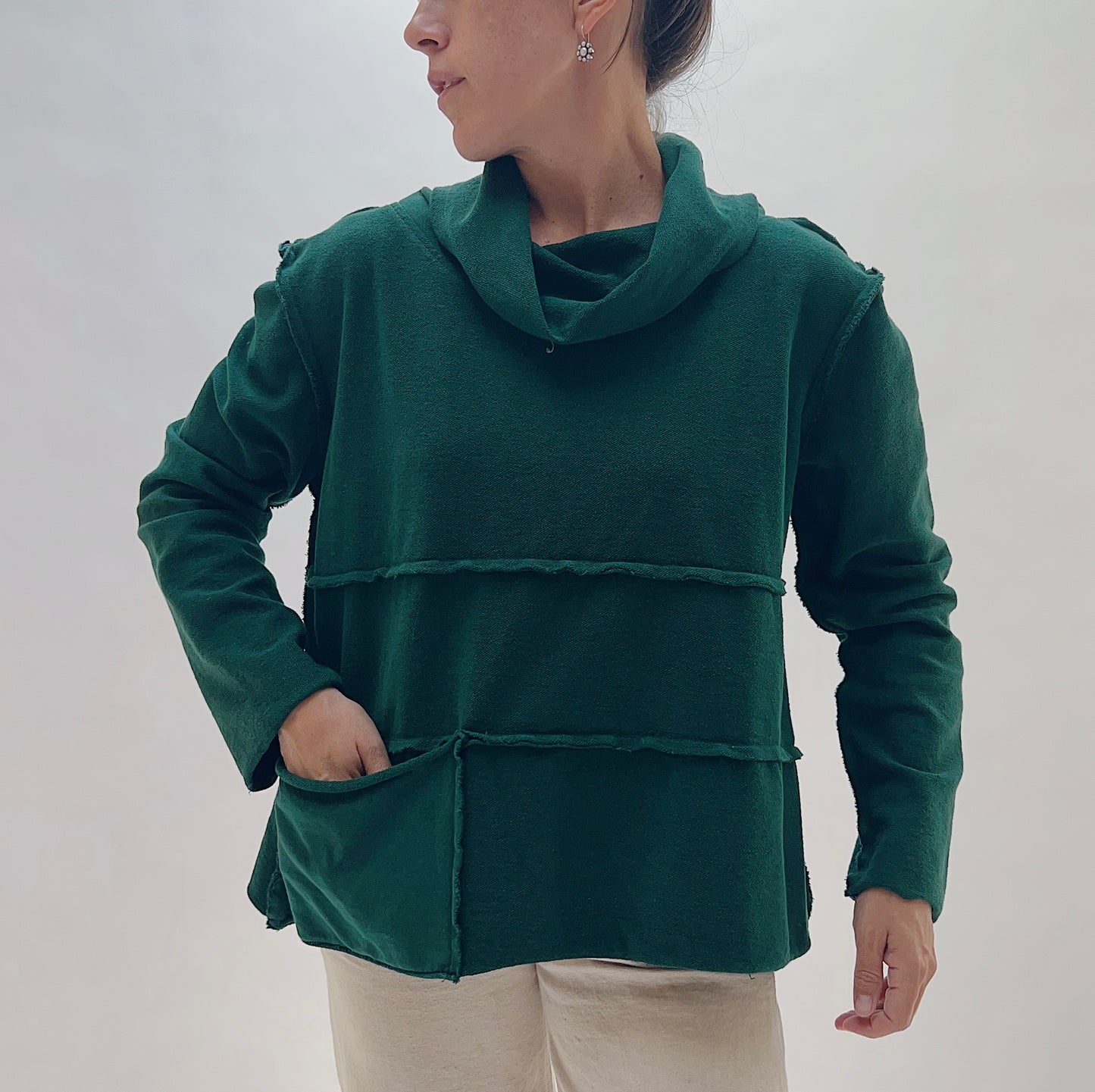 A woman wearing a dark green cowl-neck sweater with layered horizontal ruffle details and front pockets, paired with light beige pants, against a plain light gray background.
