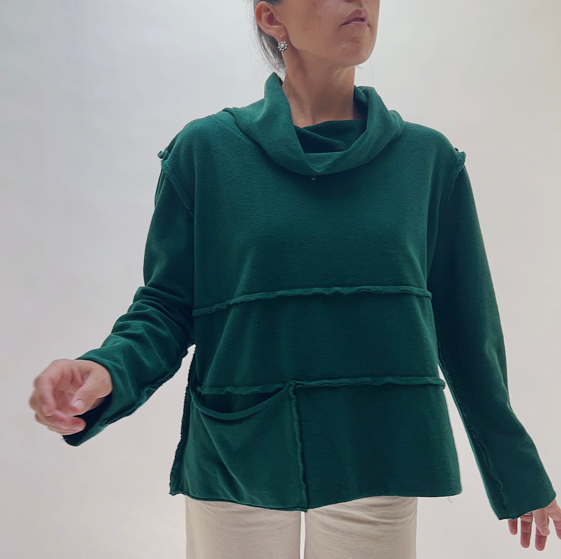 A woman wearing a dark green cowl-neck sweater with layered horizontal seams and front pockets, paired with light beige pants, against a plain light gray background.