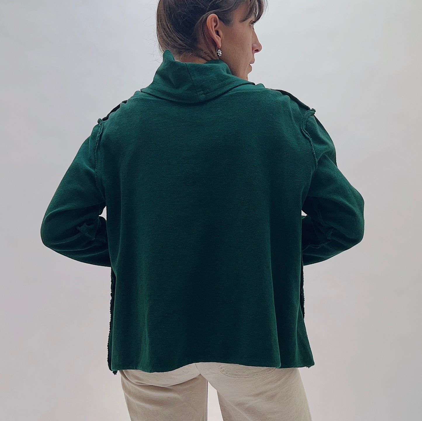 A woman shown from the back wearing a dark green long-sleeve cropped top with shoulder detailing and cream pants, set against a plain light gray background.