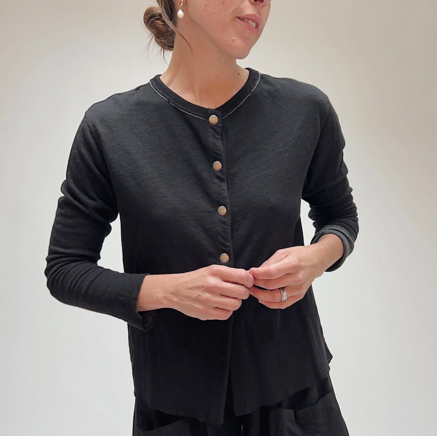 Model fastening the snap closure on the black cotton cardigan, showing crew neckline with contrast stitching detail