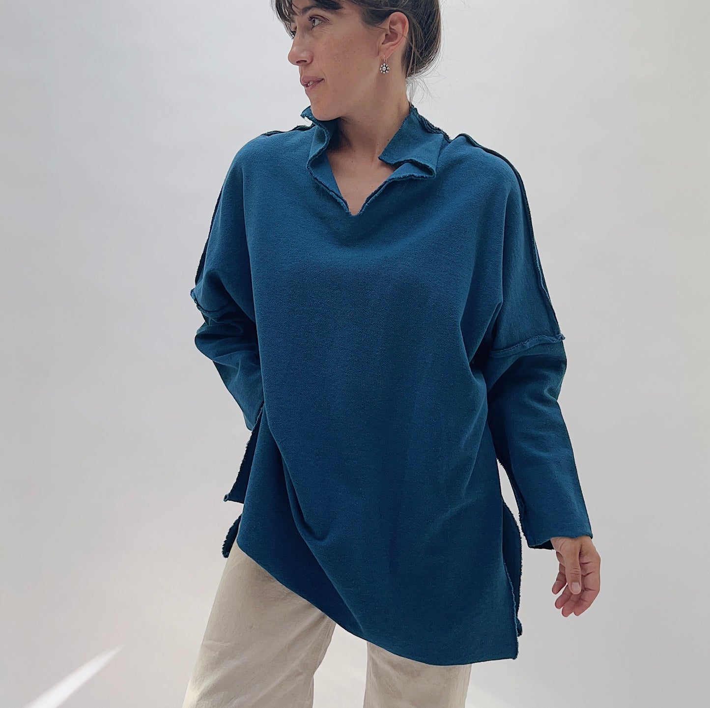 Eleven Stitch Funnel Neck Tunic in North Sea — front three-quarter view showing the open funnel collar, raw-edge exposed seams at the shoulders, dropped sleeves, and side slits in deep teal, worn with cream trousers on a white background