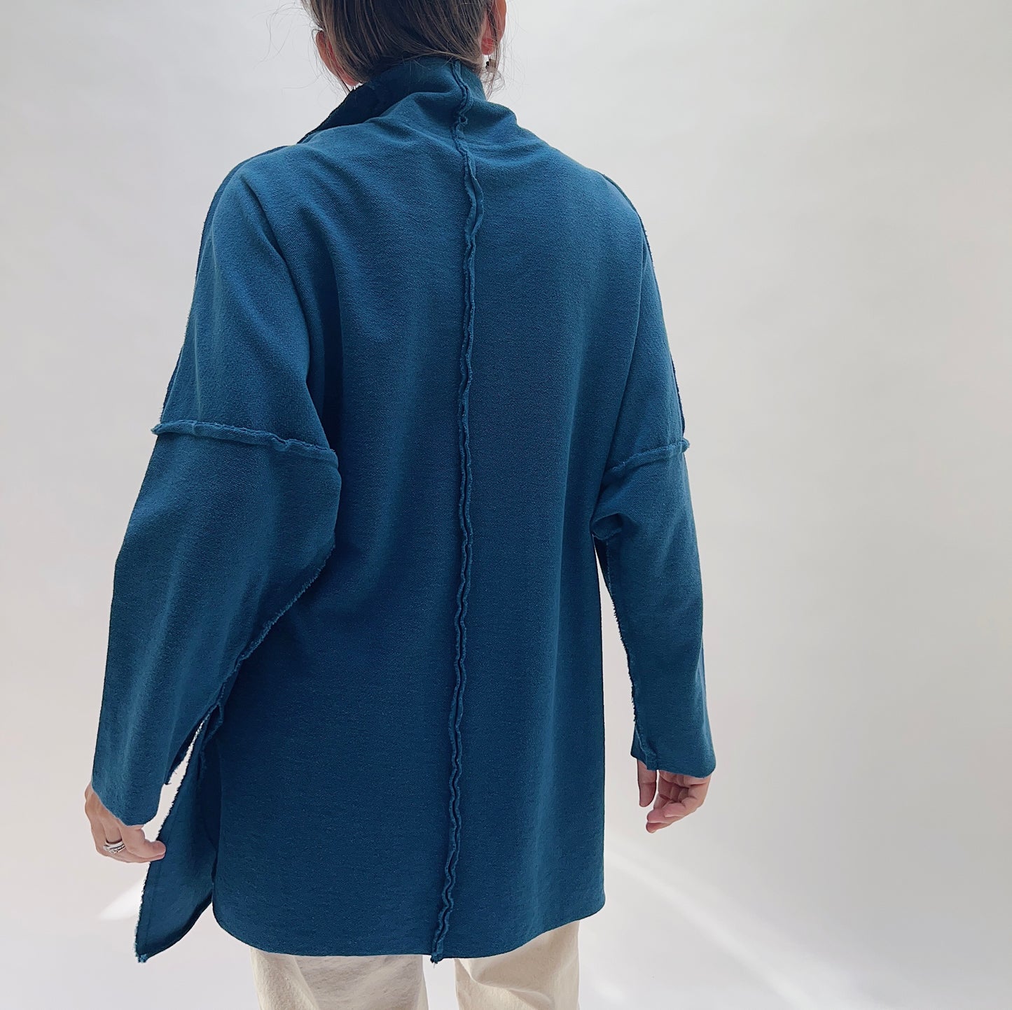 Eleven Stitch Funnel Neck Tunic in North Sea — rear view showing the center back exposed seam, raw-edge shoulder seams, wide dropped sleeves, and side slits in deep teal, worn with cream trousers on a white background