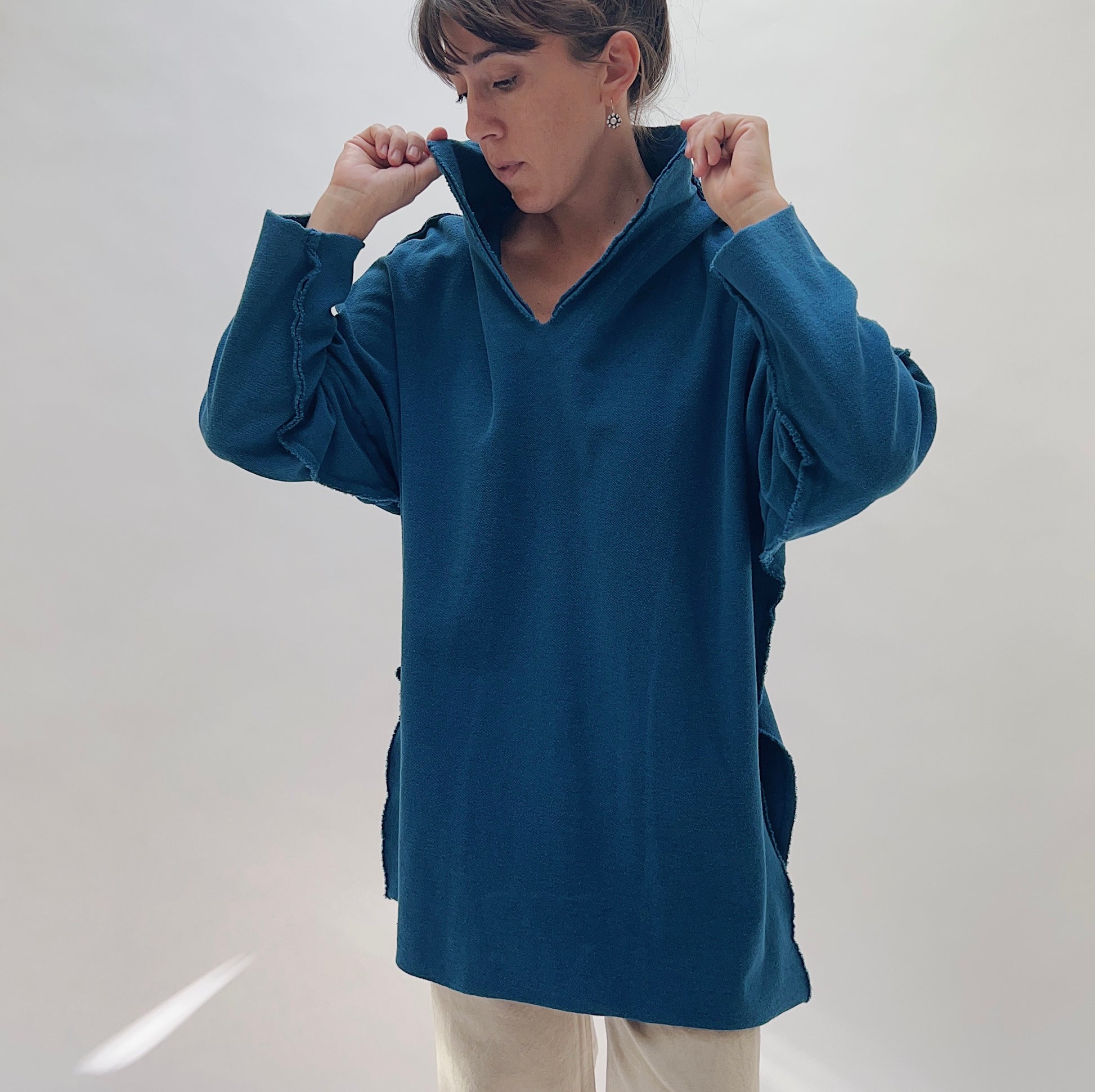 Eleven Stitch Funnel Neck Tunic in North Sea — front view with hands pulling up the open funnel collar, showing the V-notch neckline, raw-edge seam detailing, and relaxed tunic length in deep teal, worn with cream trousers on a white background