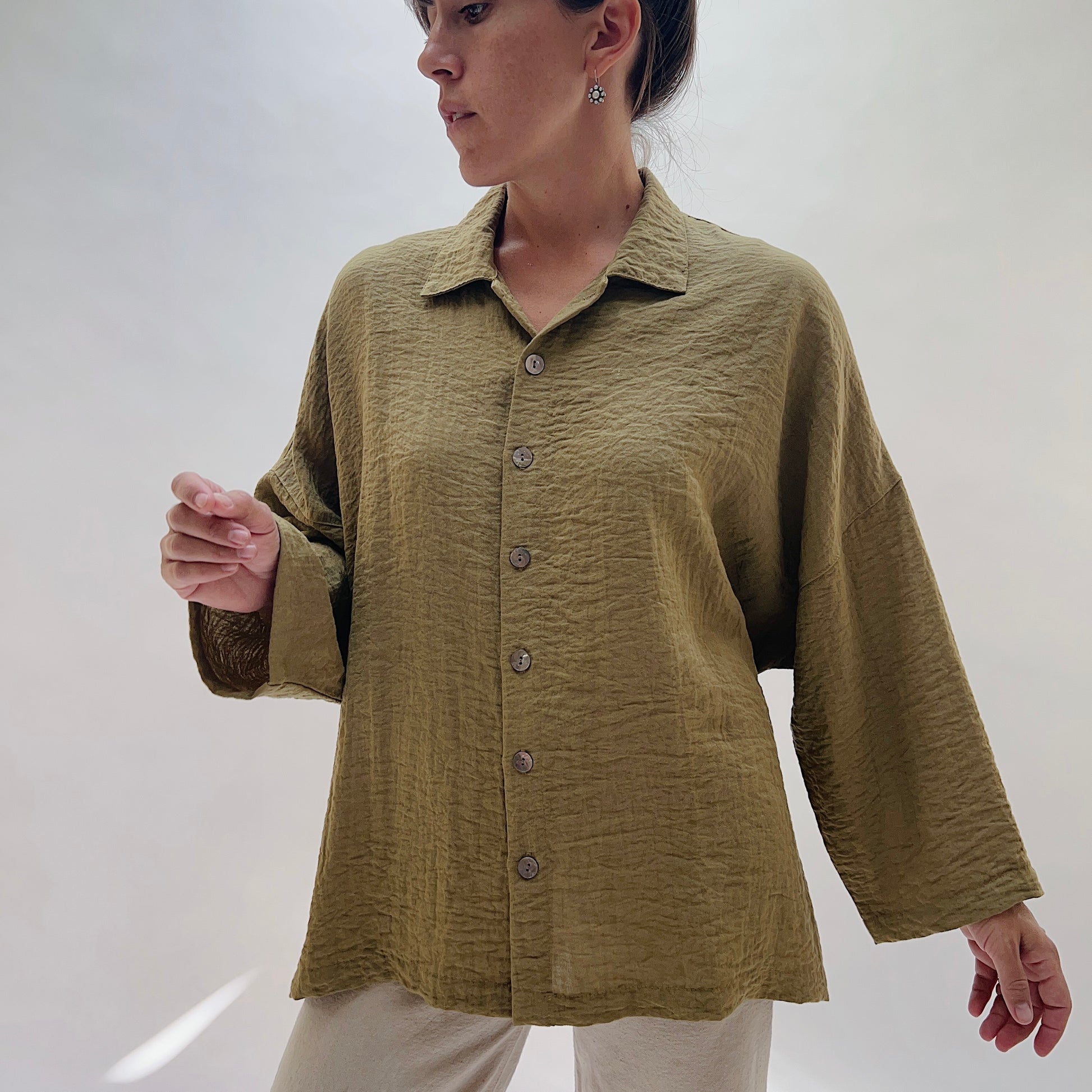 A 3/4 front view of a woman wearing a textured olive green long-sleeve button-up shirt with a relaxed fit, paired with light beige pants, photographed against a neutral light gray background.