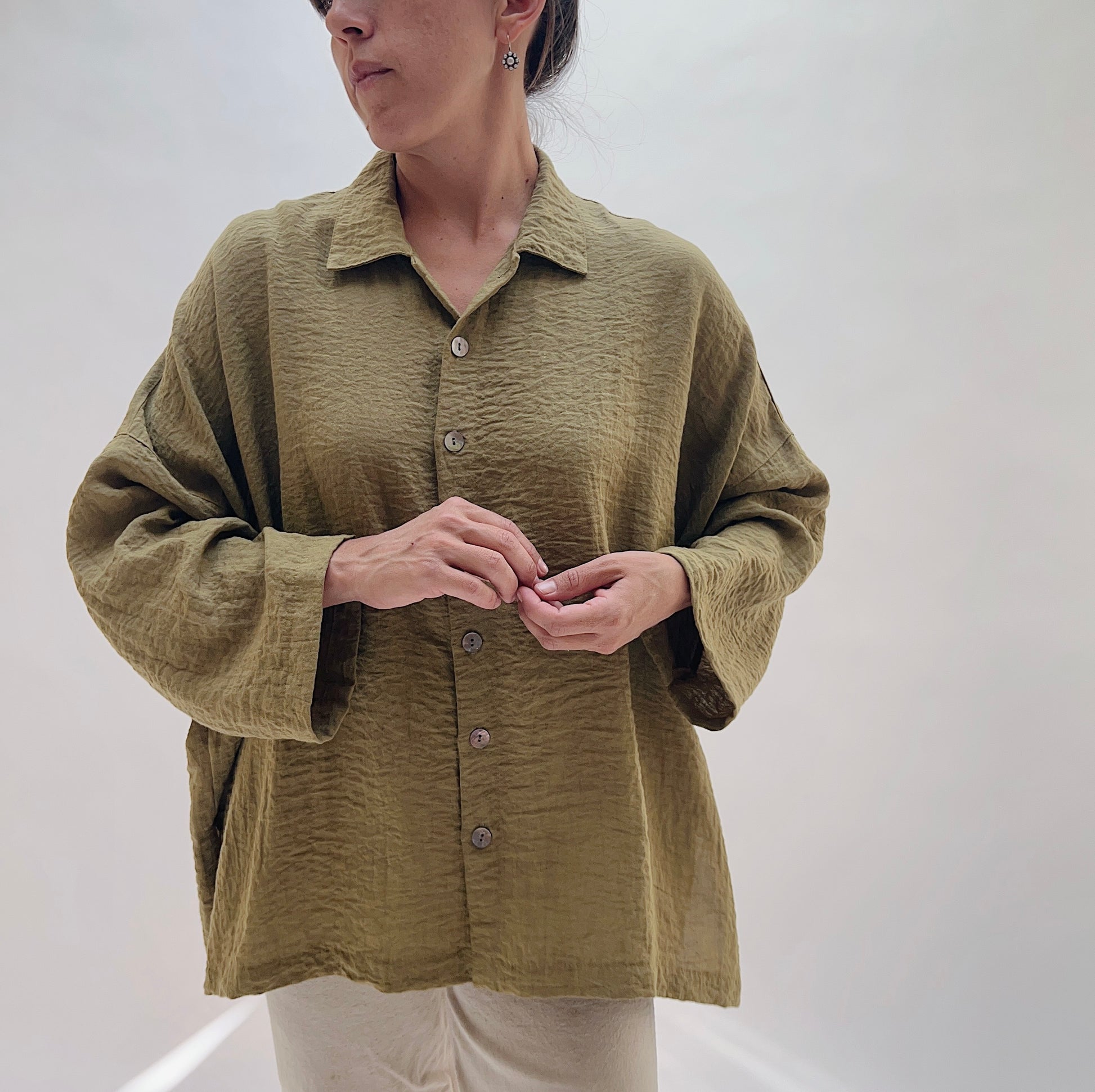 A woman wearing an oversized, textured olive green button-up shirt with wide sleeves and light beige pants, standing against a plain light gray background.