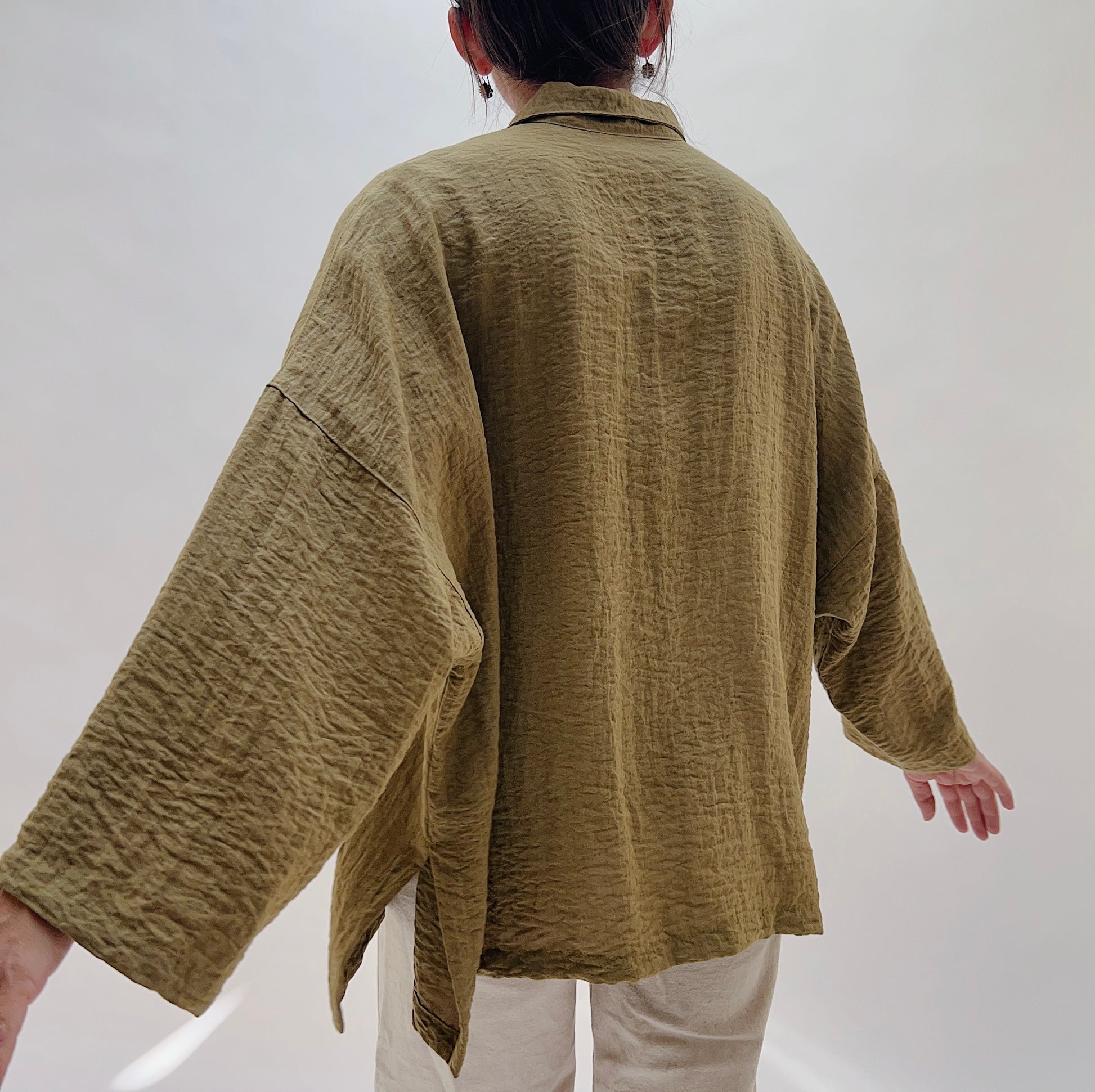 A person shown from the back wearing a loose, textured olive-brown long-sleeve shirt with wide sleeves and light beige pants, photographed against a plain light background.