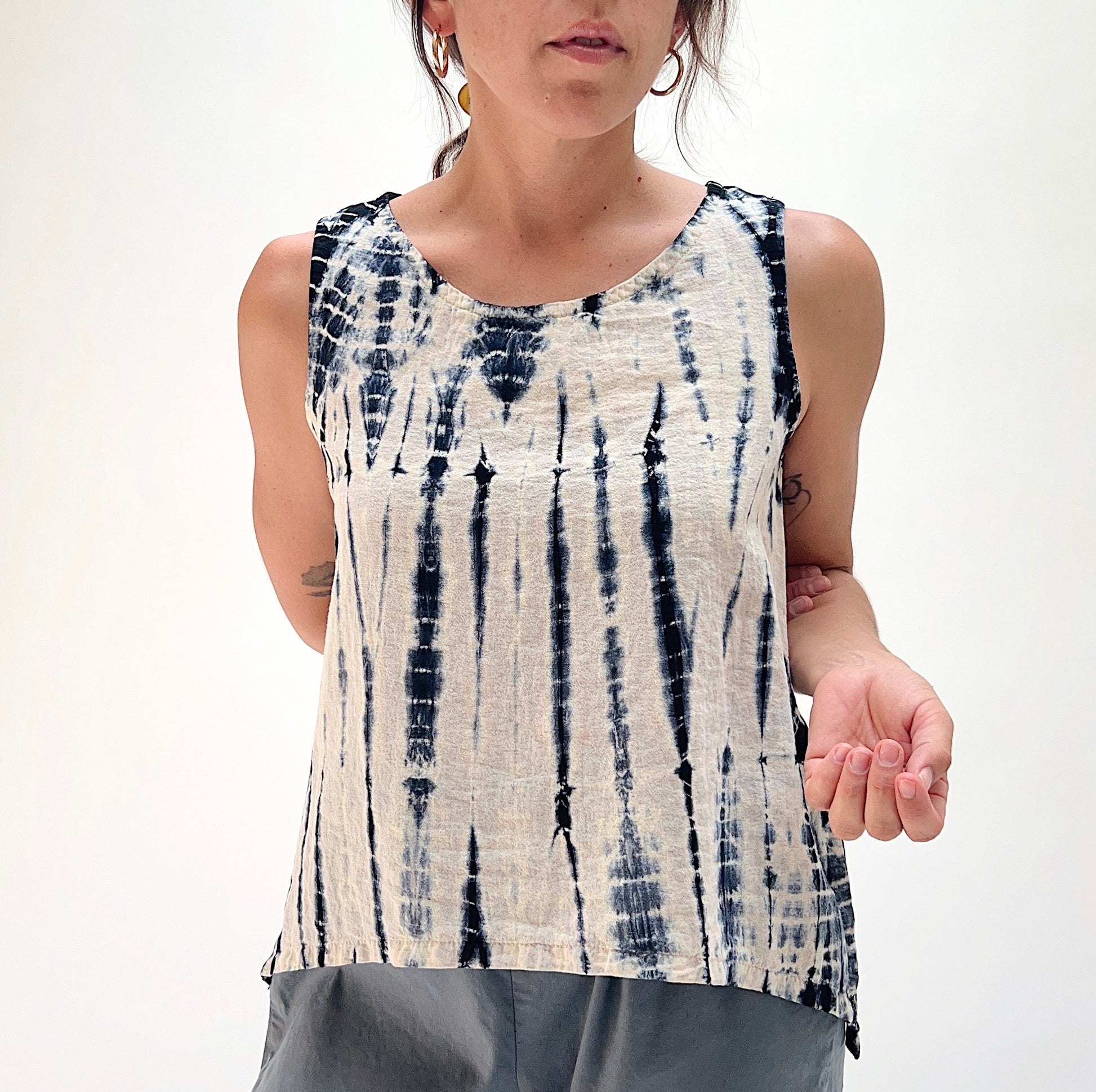 A woman wearing a sleeveless tie-dye top with a vertical indigo and cream pattern, paired with gray pants, against a plain white background.