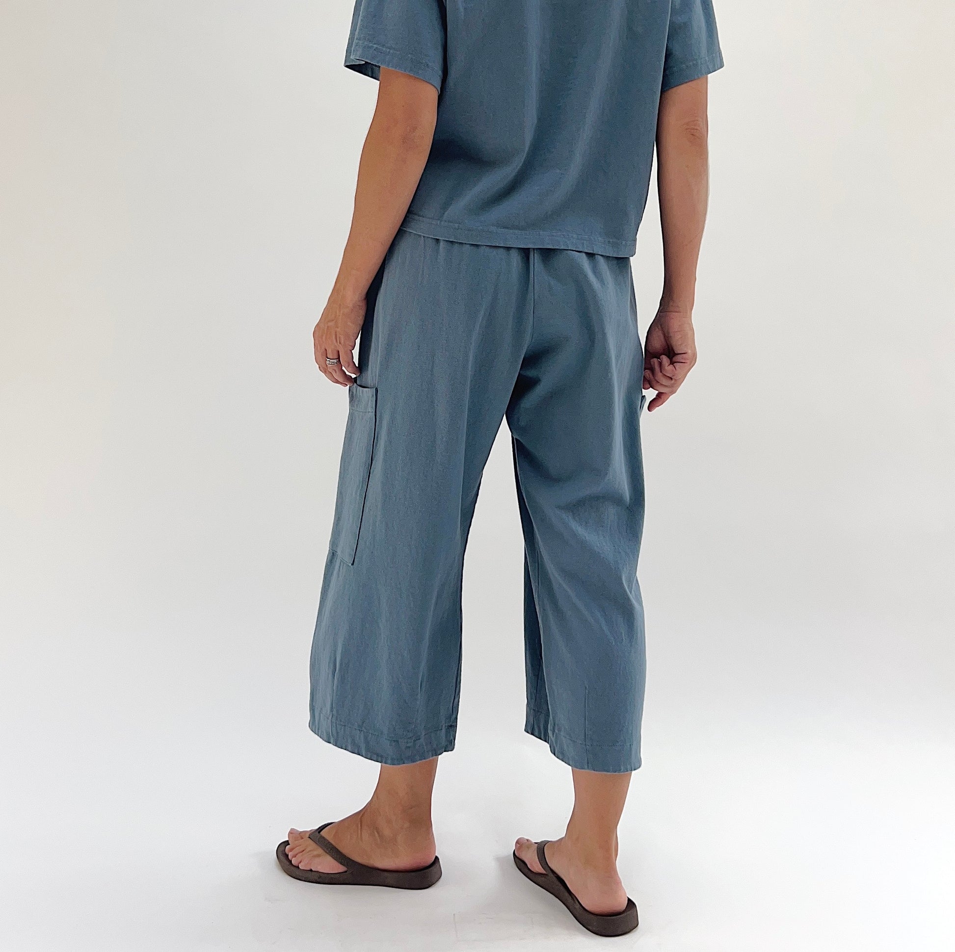 Back view of model wearing Bryn Walker vitznau cotton Pasha pant, showing large patch pocket on back leg, wide cropped silhouette, elastic waistband, and dusty blue color
