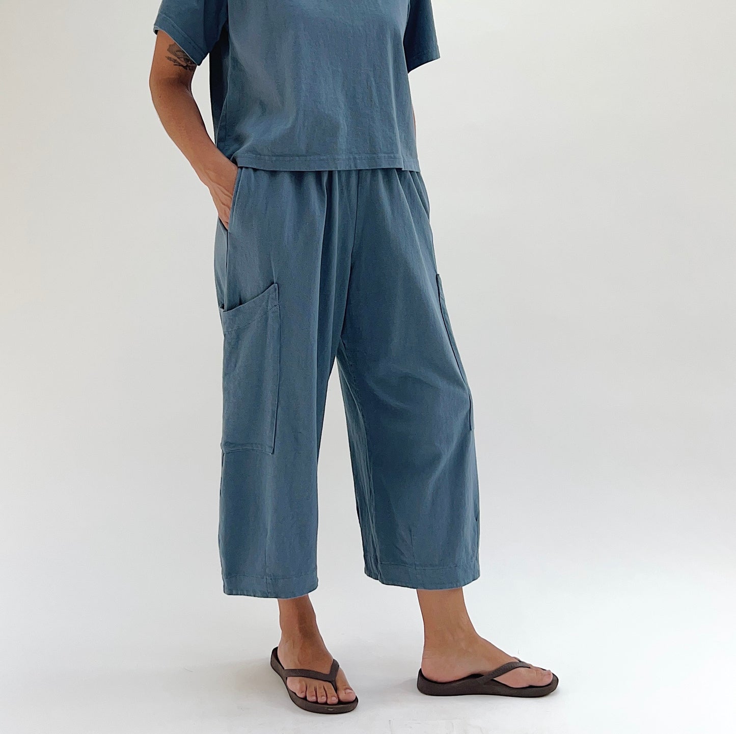 Front three-quarter view of model wearing Bryn Walker vitznau cotton Pasha pant, showing large patch pockets on each leg, elastic waistband, wide cropped leg, and dusty blue color