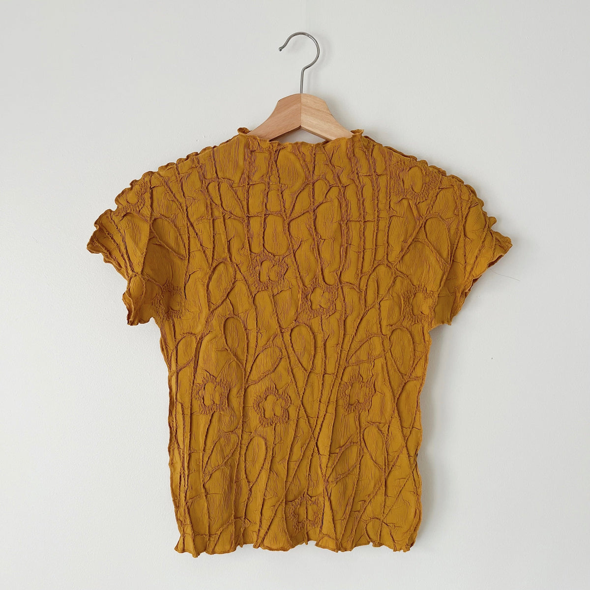 Front view of the mustard short sleeve crinkle top on a wooden hanger, showing its raised textured pattern.
