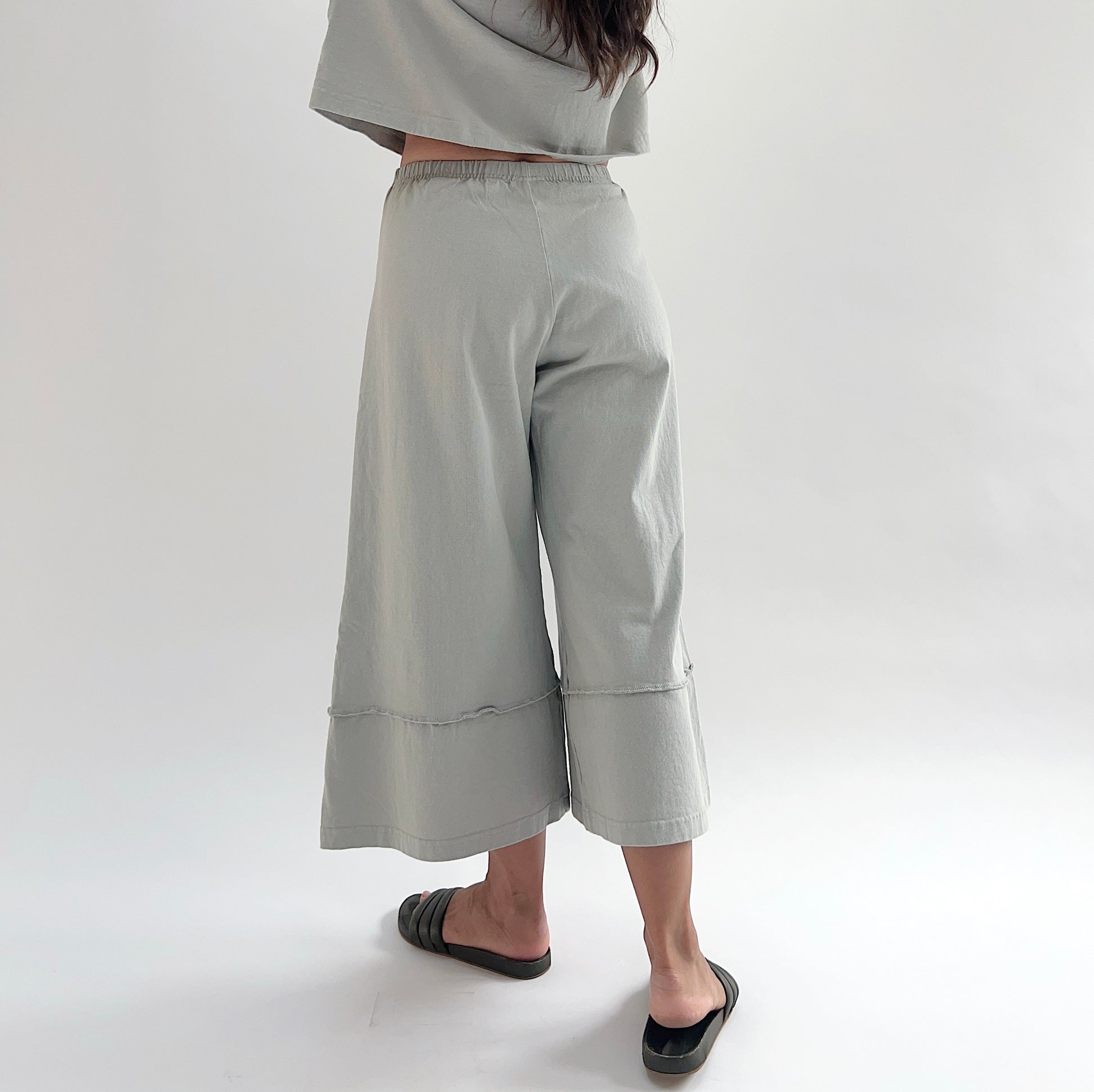 Back three-quarter view of model wearing Pacific Cotton orlaya crop full pant, showing elastic waistband, wide flared leg, layered hem panel, and sage green color