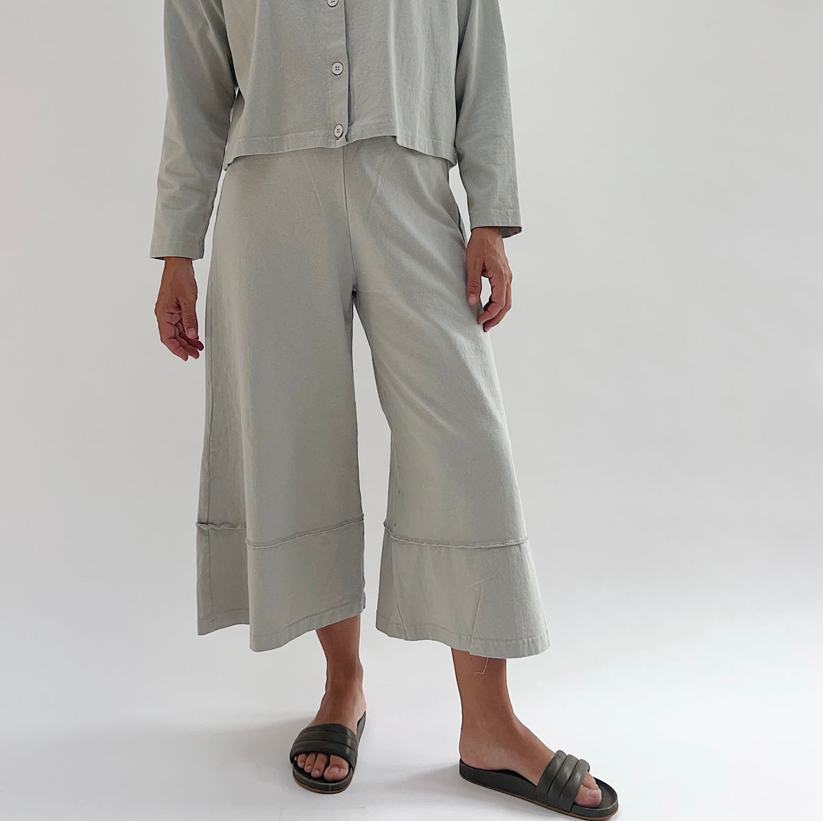 Full front view of model wearing Pacific Cotton orlaya crop full pant, showing elastic waistband, wide flared leg, layered hem panel, and cropped length in sage green