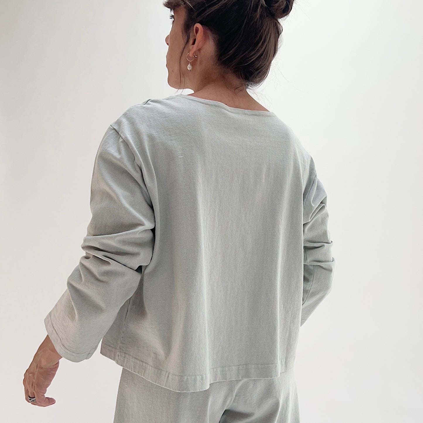 Back view of model wearing Pacific Cotton orlaya boxy shirt, showing long sleeves, relaxed boxy cropped silhouette, banded hem, and sage green color