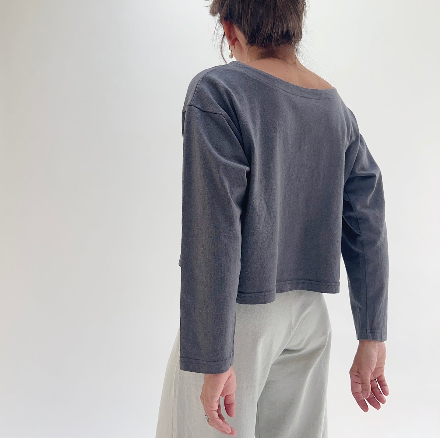 Back view of model wearing Pacific Cotton moonlight long sleeve button front shirt, showing wide back neckline, long sleeves, boxy cropped silhouette, banded hem, and slate blue-grey color