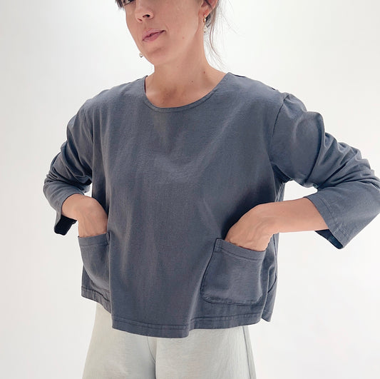 A woman wearing a dark slate gray long-sleeve top with large front patch pockets, paired with light gray pants, photographed against a clean white studio background.