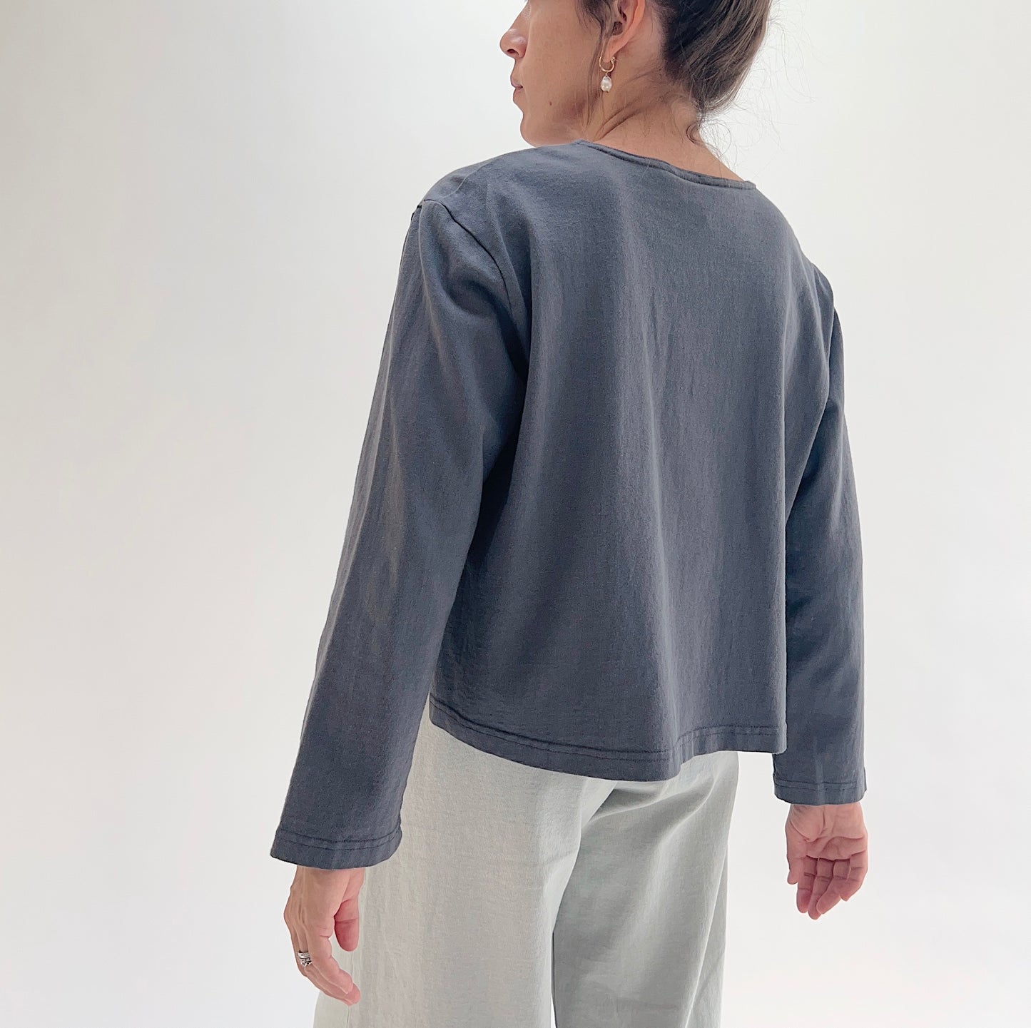 A woman shown from the back wearing a dark slate gray long-sleeve cropped top and light gray pants, photographed against a clean white studio background.