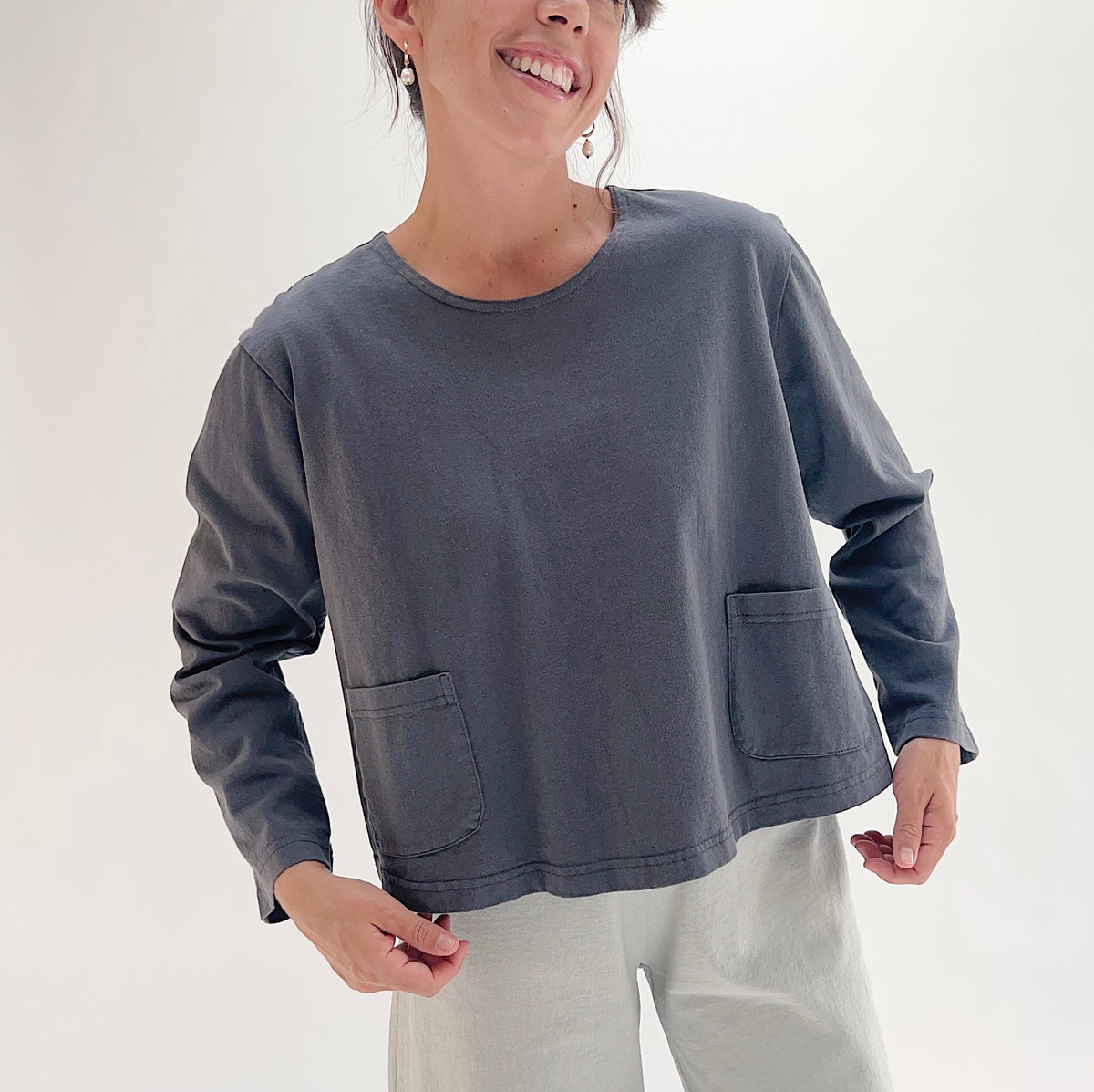 A woman wearing a dark gray long-sleeve top with two front patch pockets, paired with light gray pants, photographed against a plain white studio background.