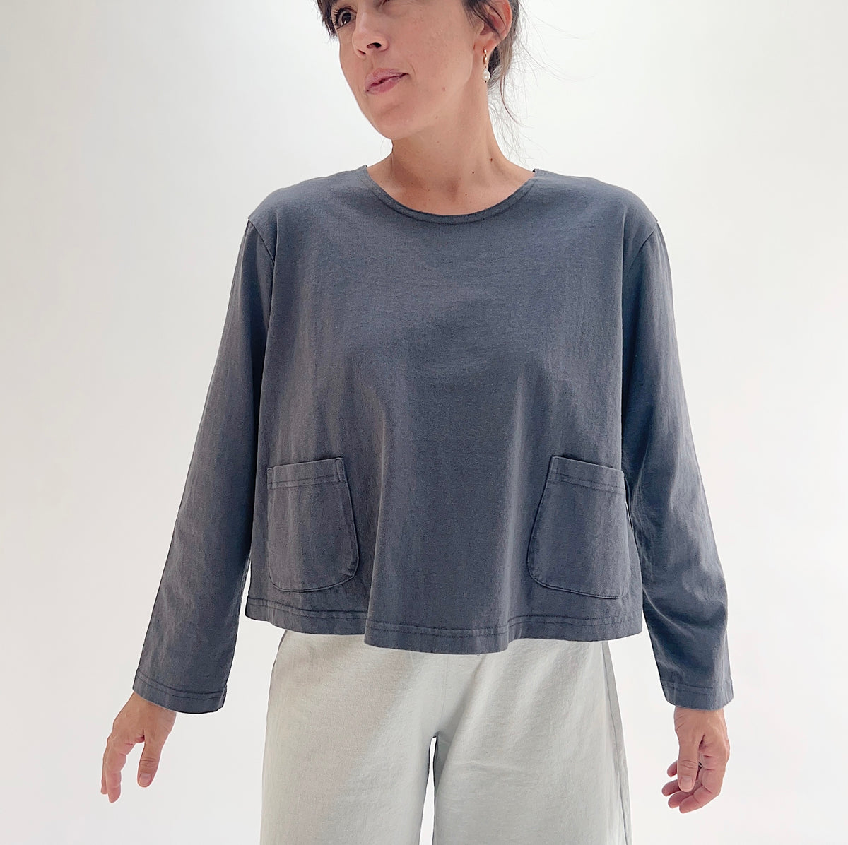 Three-quarter front view of model wearing Pacific Cotton moonlight boxy shirt, showing crew neckline, two front patch pockets, long sleeves, relaxed boxy cropped silhouette, and slate blue-grey color