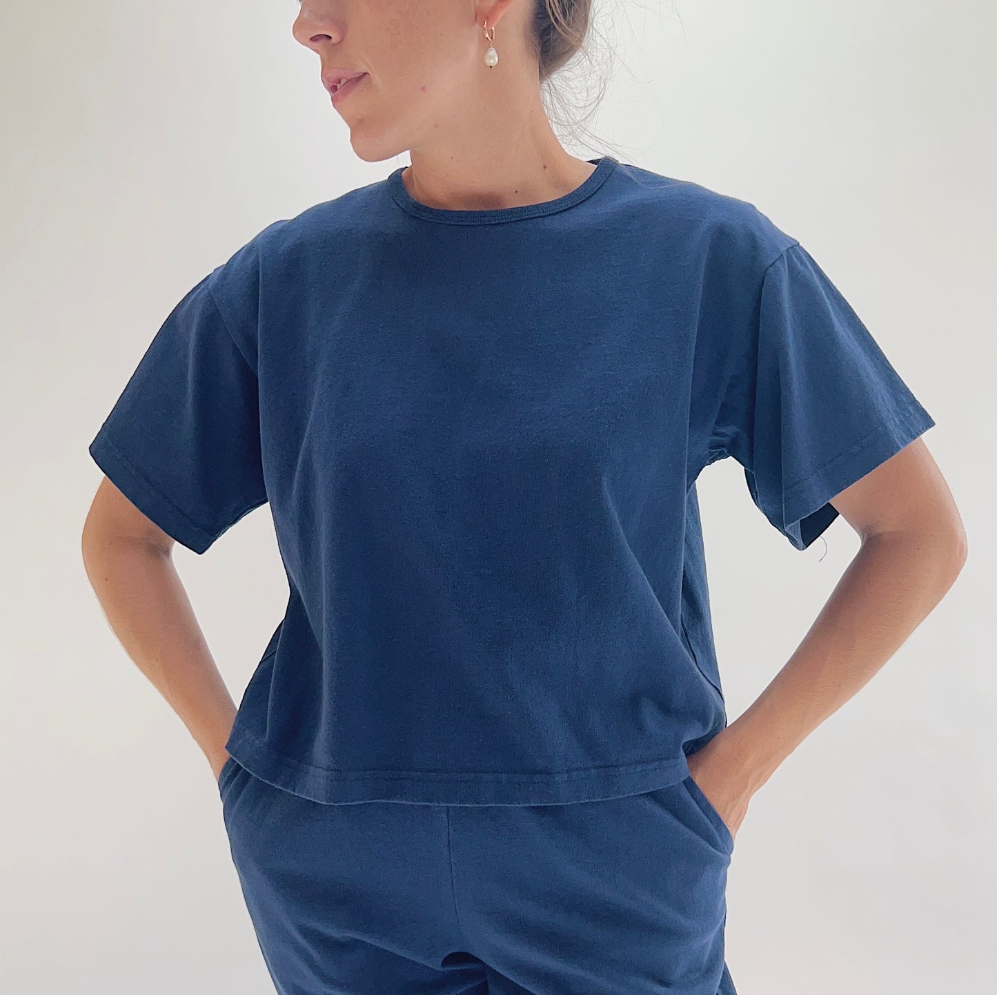 Close front view of model wearing Pacific Cotton nova crop crew tee with hands on hips, showing crew neckline, short sleeves, boxy cropped fit, banded hem, and deep teal color