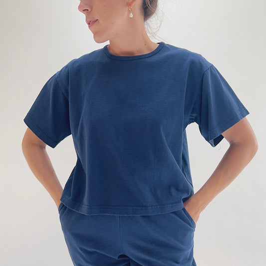 Close front view of model wearing Pacific Cotton nova crop crew tee with hands on hips, showing crew neckline, short sleeves, boxy cropped fit, banded hem, and deep teal color
