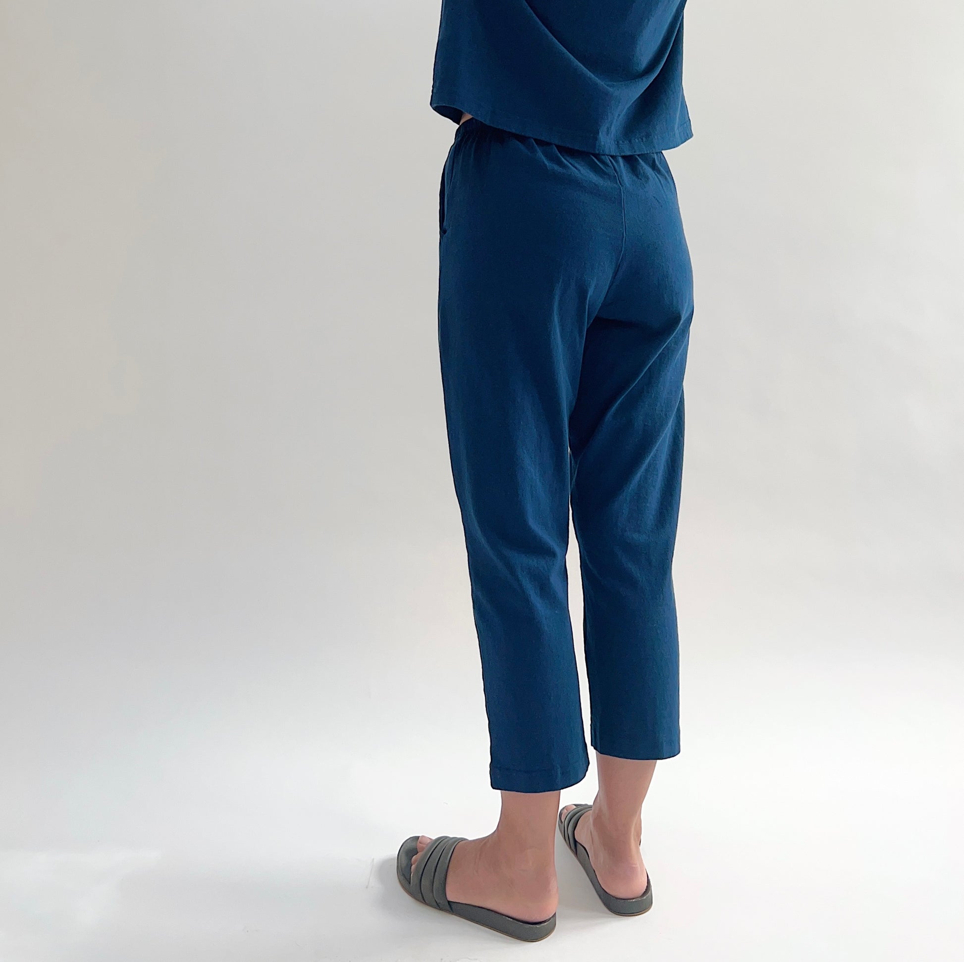 Three-quarter back view of model wearing Pacific Cotton nova sunday pant, showing elastic waistband, side pocket, straight cropped leg, and deep teal color, styled with matching nova crop crew tee