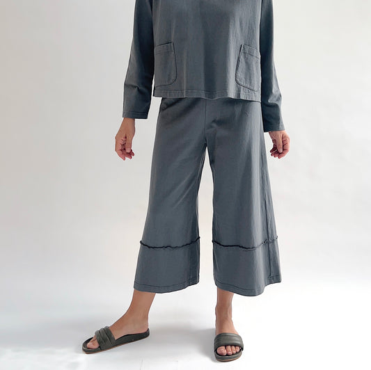 Close front view of model wearing Pacific Cotton dolomite crop full pant, showing elastic waistband, wide flared leg, layered hem panel with exposed seam detail, and washed slate grey color, styled with matching dolomite boxy shirt