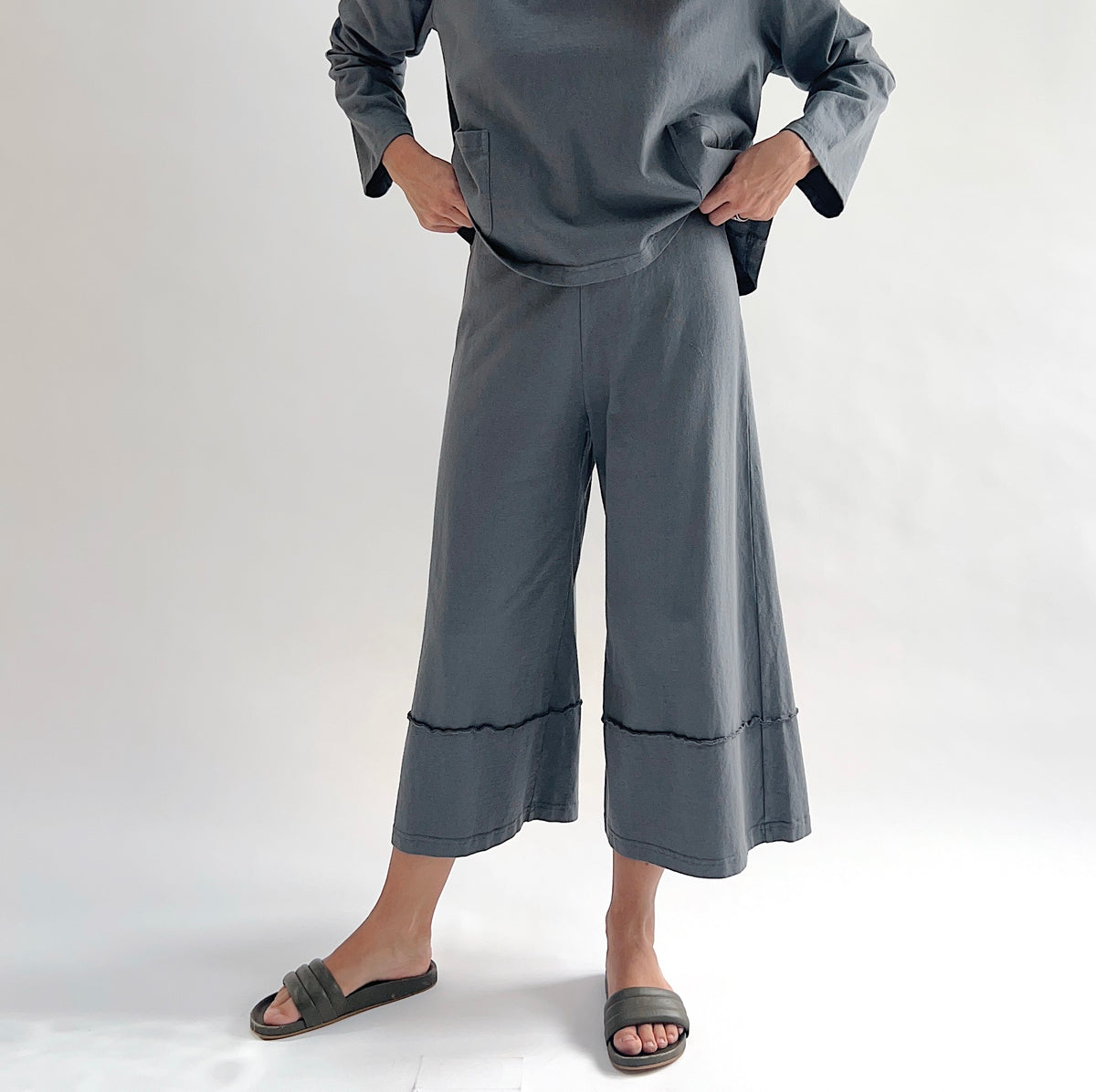 Full front view of model wearing Pacific Cotton dolomite crop full pant with hands on hips, showing elastic waistband, wide flared leg silhouette, layered hem panel, and washed slate grey color, styled with matching dolomite boxy shirt