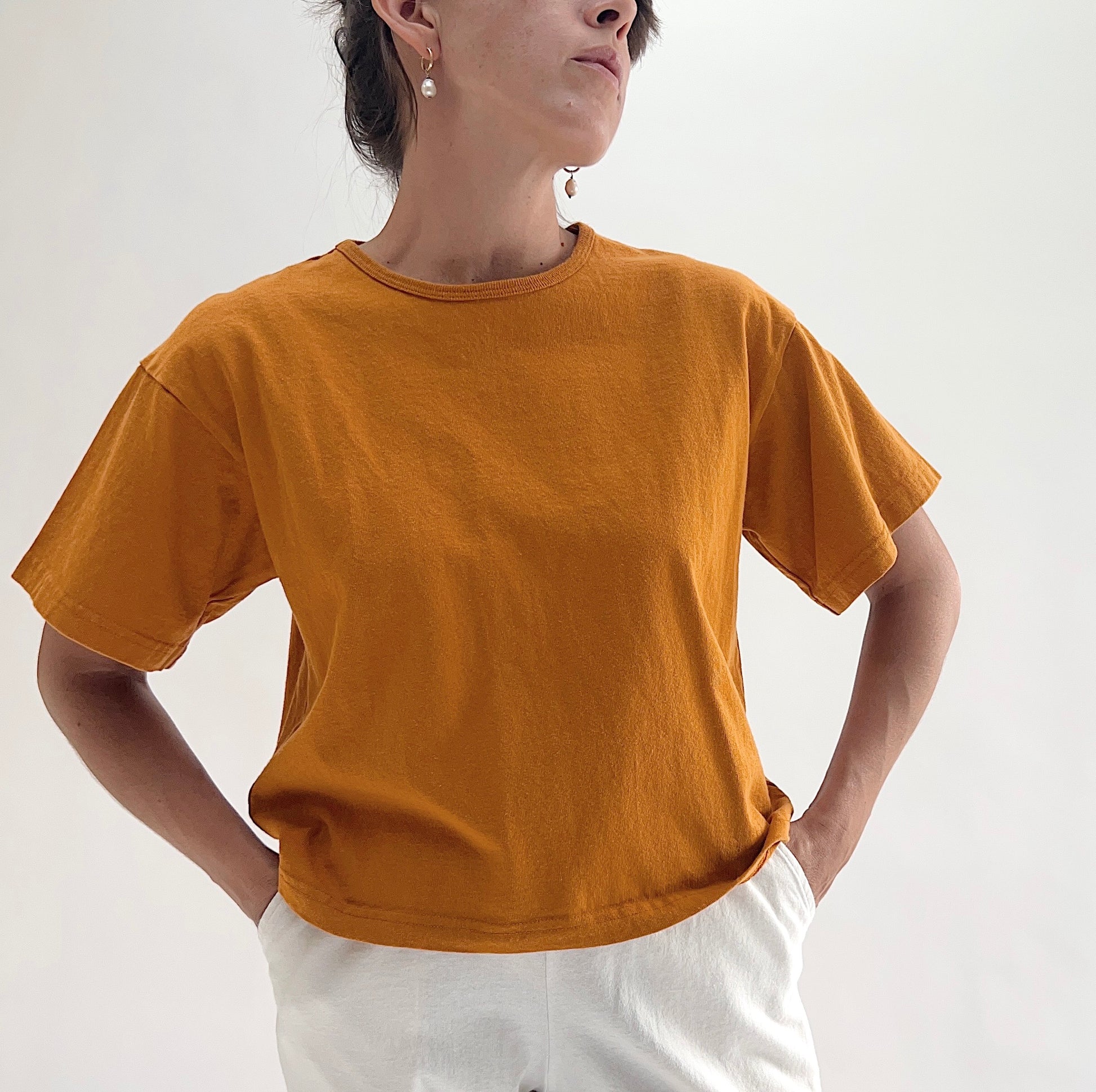 Close front view of model wearing Pacific Cotton yera crop crew tee with hands on hips, showing ribbed crew neckline, short cuffed sleeves, boxy cropped fit, and warm golden amber color