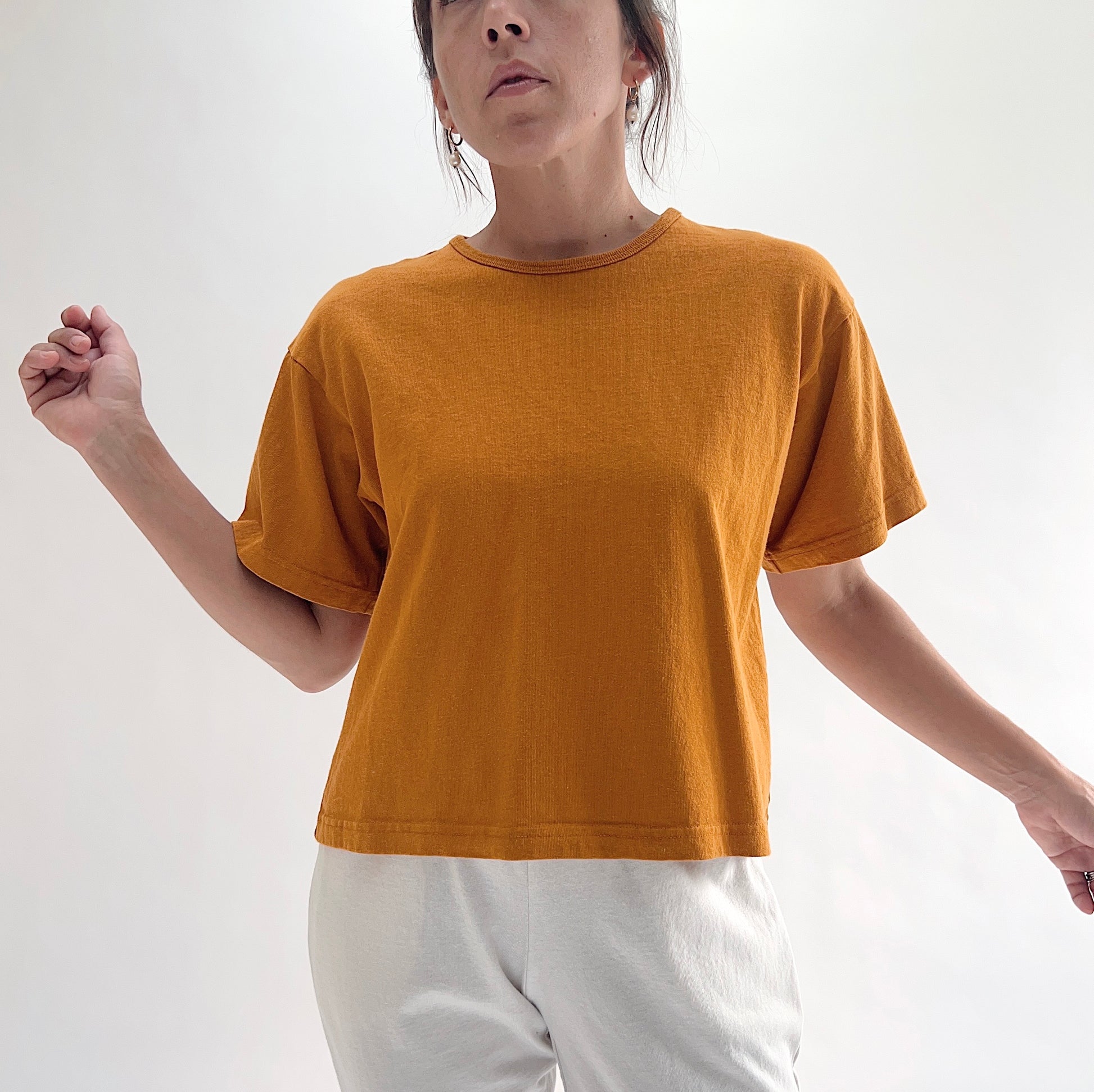 Full front view of model wearing Pacific Cotton yera crop crew tee, showing ribbed crew neckline, short cuffed sleeves, boxy cropped silhouette, banded hem, and warm golden amber color