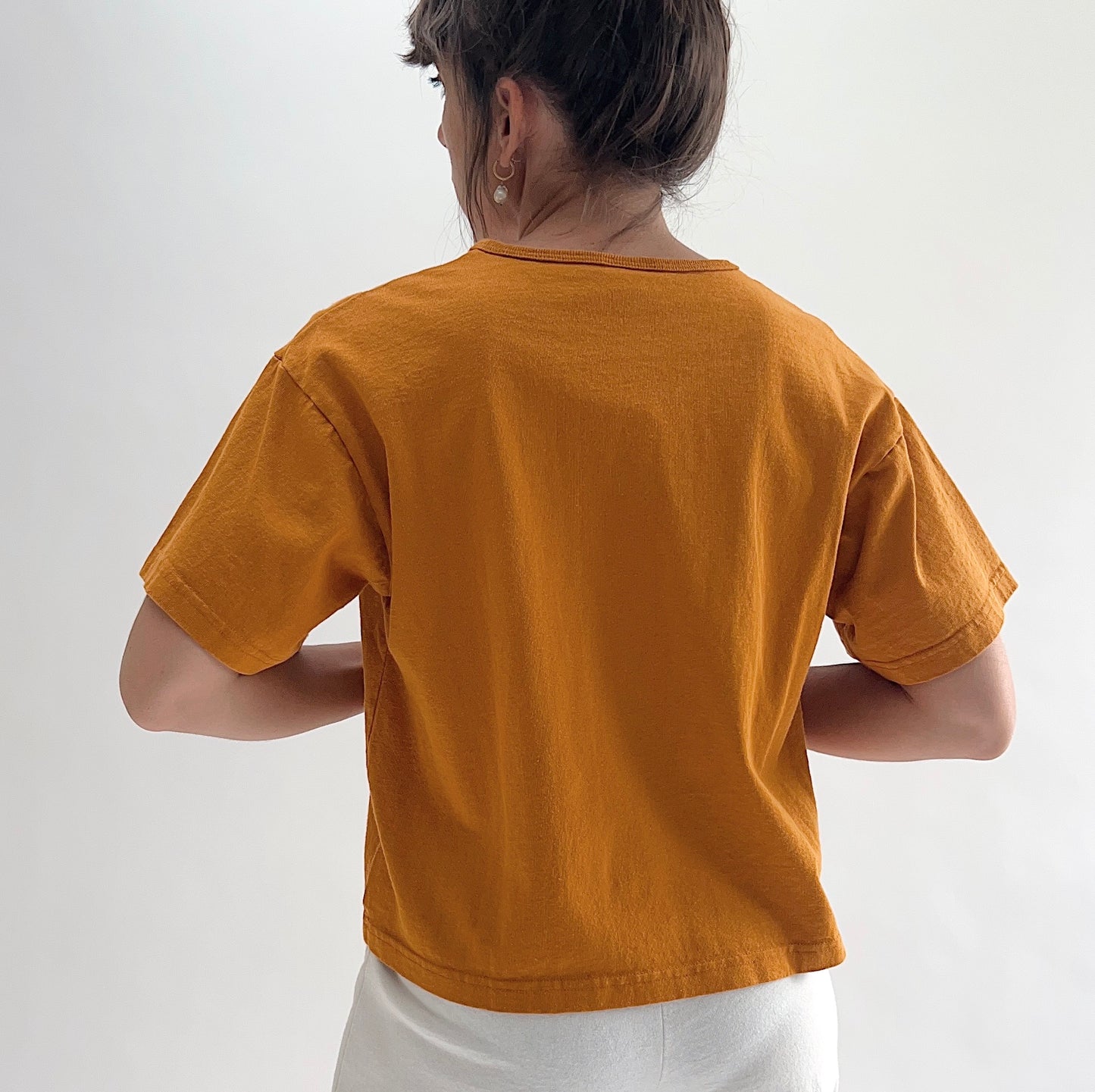 Back view of model wearing Pacific Cotton yera crop crew tee, showing crew neckline, short cuffed sleeves, boxy cropped silhouette, banded hem, and warm golden amber color