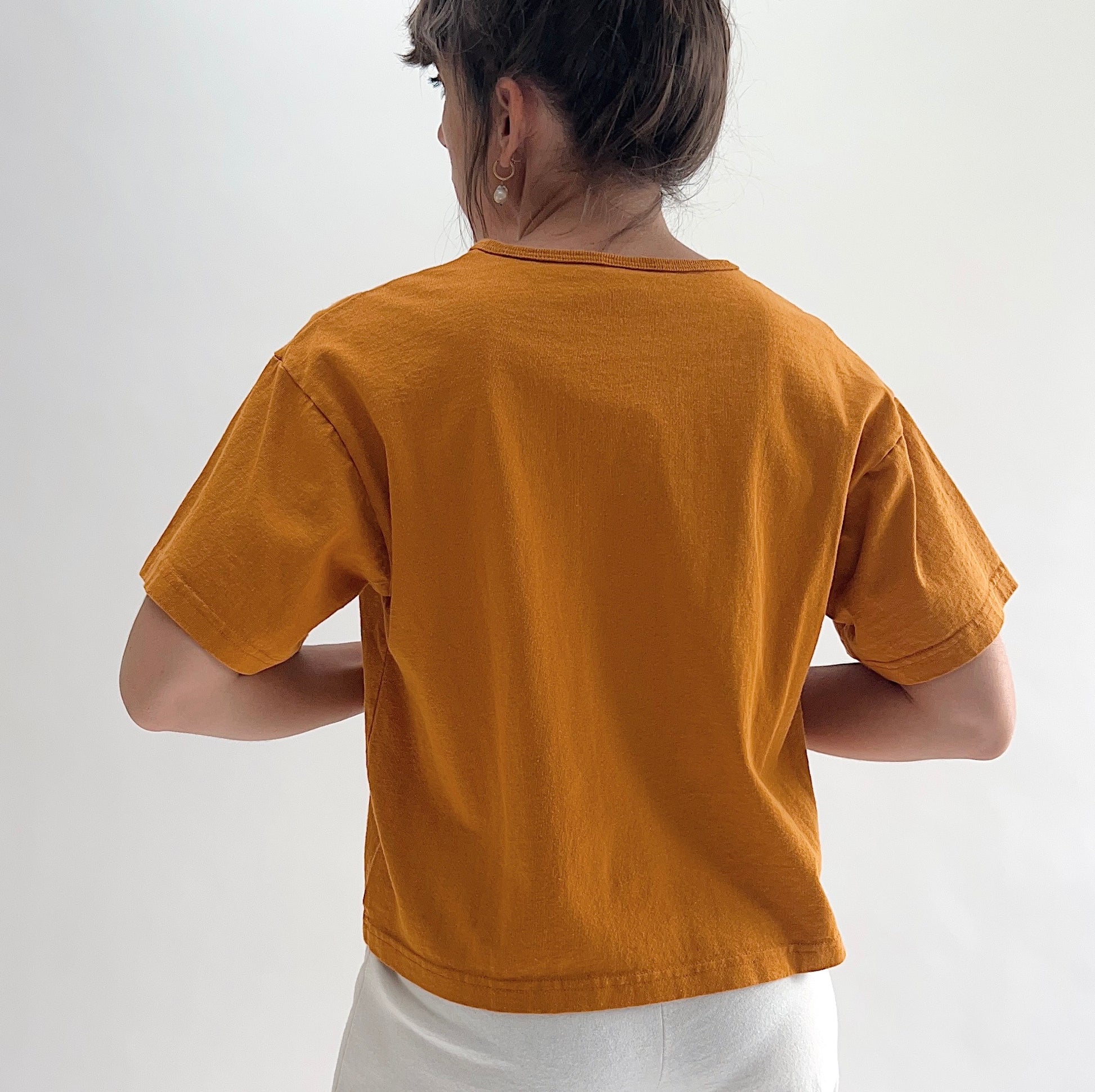 Back view of model wearing Pacific Cotton yera crop crew tee, showing crew neckline, short cuffed sleeves, boxy cropped silhouette, banded hem, and warm golden amber color