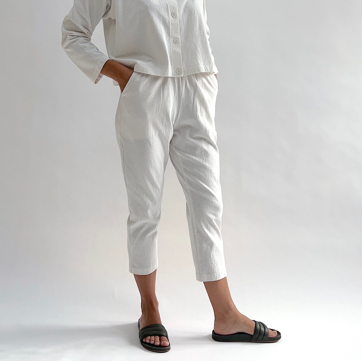 Close front view of model wearing Pacific Cotton cream sunday pant with hand in pocket, showing elastic waistband, side pocket, cropped tapered leg, and off-white color, styled with matching cream long sleeve button front shirt