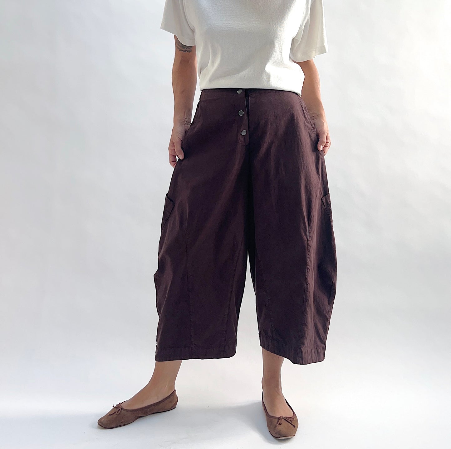 Full front view of model wearing Eleven Stitch chicory snap front pant, showing snap front waistband, wide balloon leg, large side patch pockets, gathered elastic ankle hem, and deep espresso brown color, styled with cream short sleeve tee and taupe ballet flats