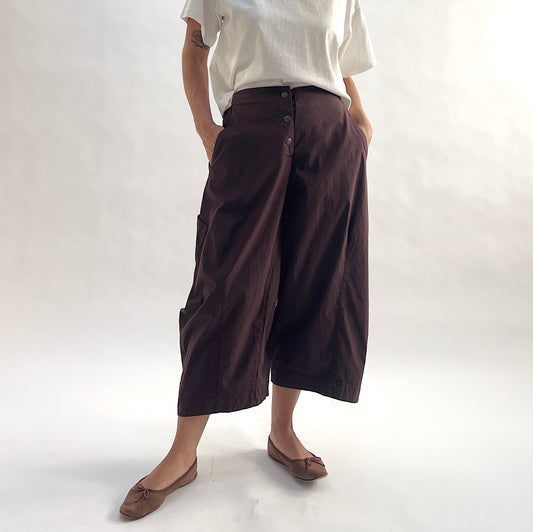 Three-quarter front view of model wearing Eleven Stitch chicory snap front pant with hands in pockets, showing snap front waistband, wide balloon leg silhouette, large side patch pockets, gathered elastic ankle hem, and deep espresso brown color, styled with cream short sleeve tee and taupe ballet flats
