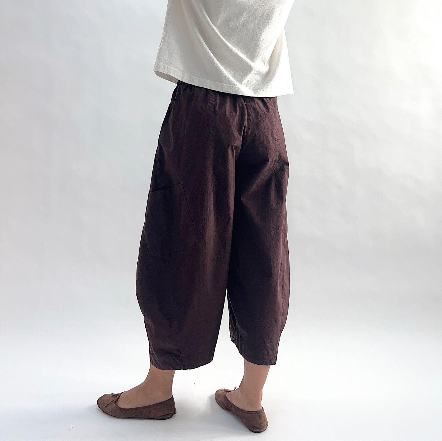 Back side view of model wearing Eleven Stitch chicory snap front pant, showing elastic back waistband, wide balloon leg, gathered elastic ankle hem, and deep espresso brown color, styled with cream short sleeve tee and taupe ballet flats
