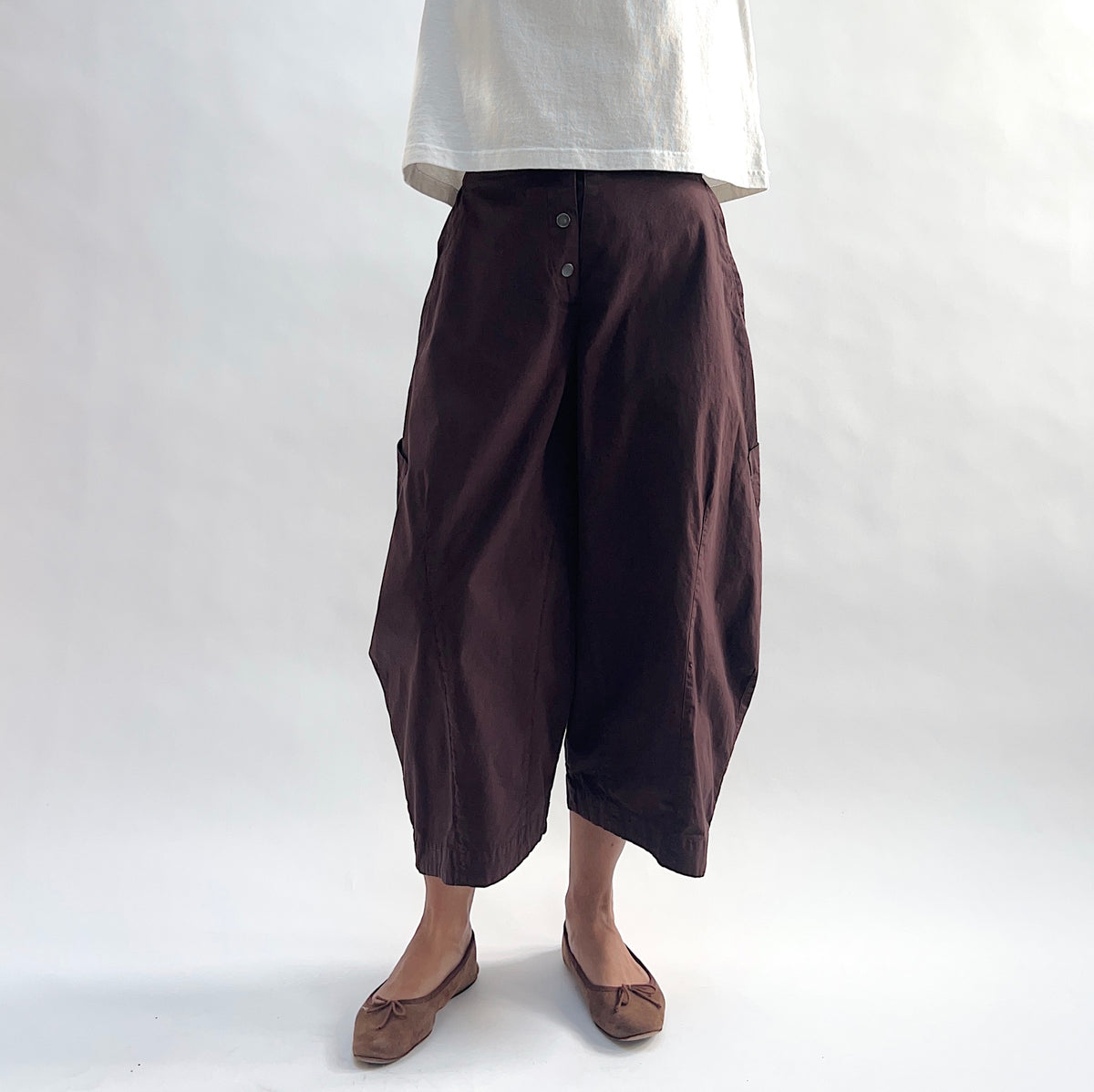 Close front view of model wearing Eleven Stitch chicory snap front pant, showing snap front waistband, wide balloon leg, large side patch pockets, gathered elastic ankle hem, and deep espresso brown color, styled with cream short sleeve tee and taupe ballet flats