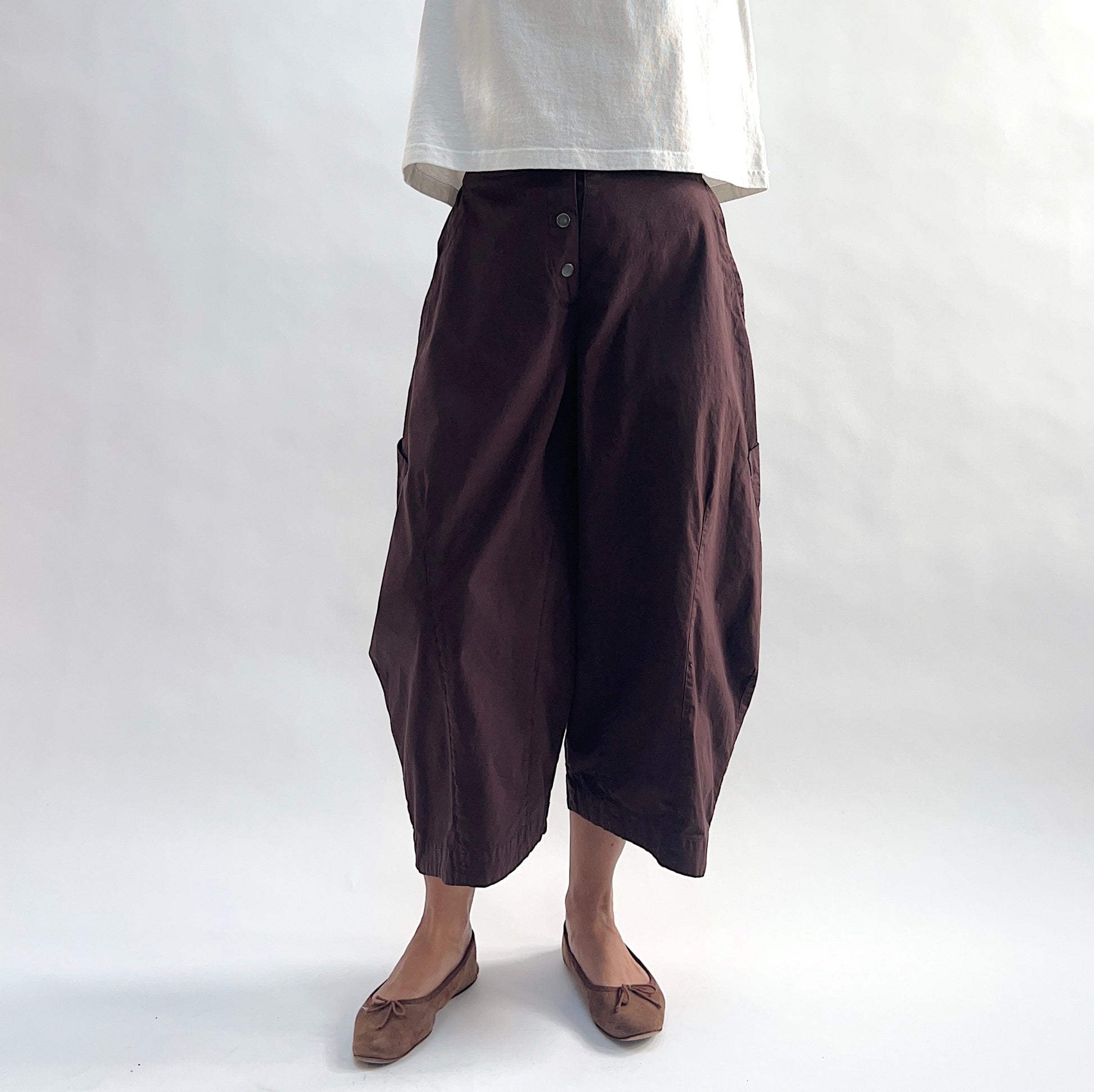Close front view of model wearing Eleven Stitch chicory snap front pant, showing snap front waistband, wide balloon leg, large side patch pockets, gathered elastic ankle hem, and deep espresso brown color, styled with cream short sleeve tee and taupe ballet flats