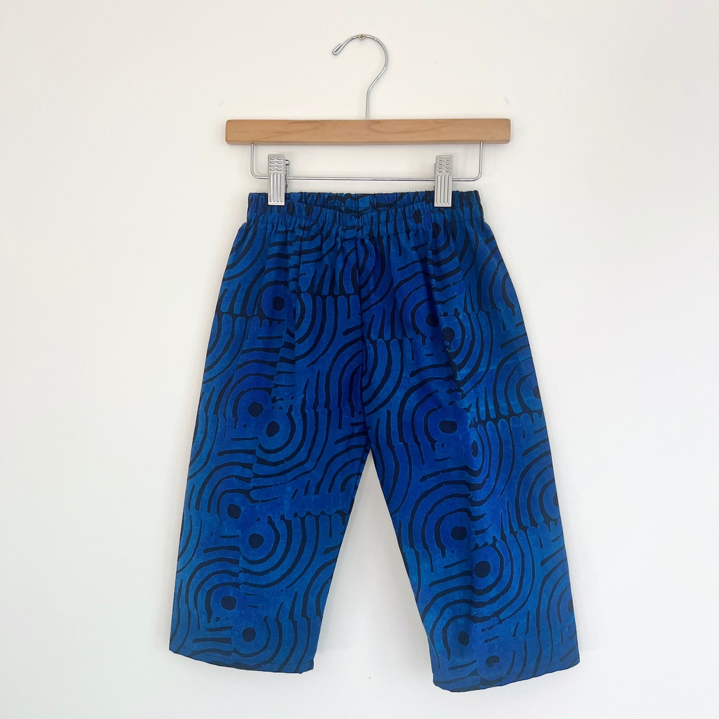 Kids lazy pants in blue waves Batiki cotton, hanging flat on a wooden clip hanger against a white wall, showing elasticated waistband, bold blue and black swirling wave print, and relaxed fit