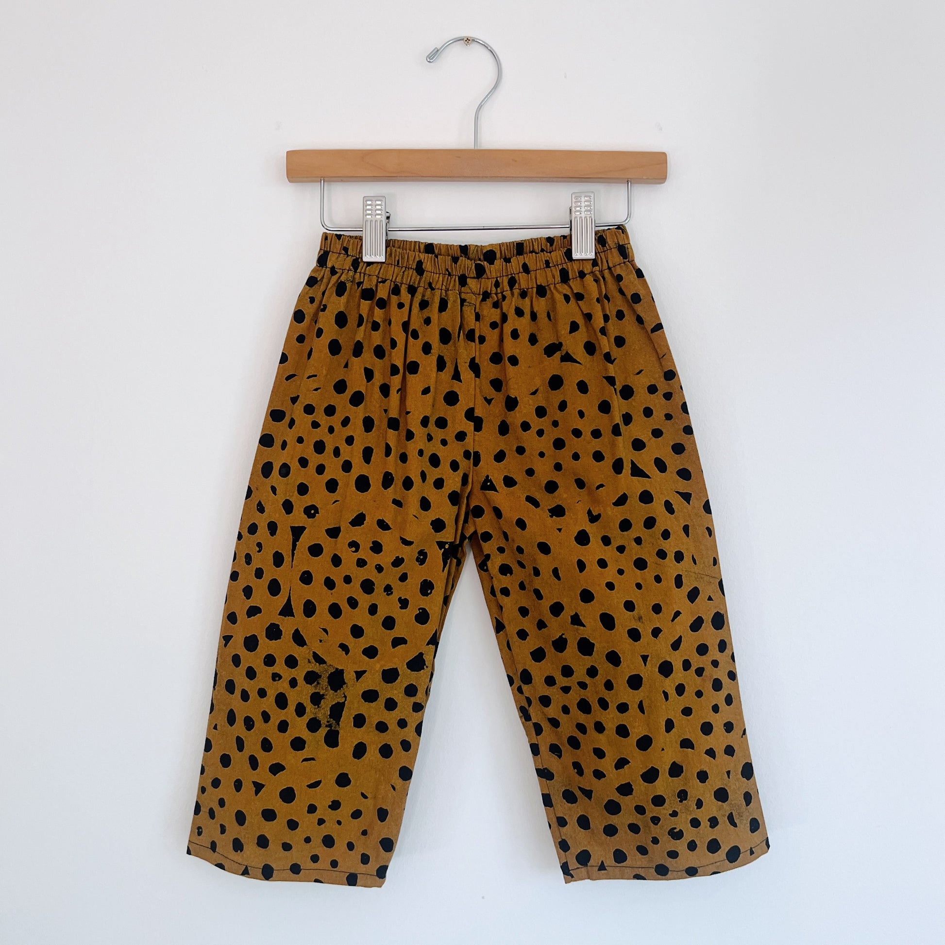 Kids lazy pants in cheetah spot Batiki cotton, hanging flat on a wooden clip hanger against a white wall, showing elasticated waistband, golden amber base with hand-printed black cheetah spots, and relaxed fit