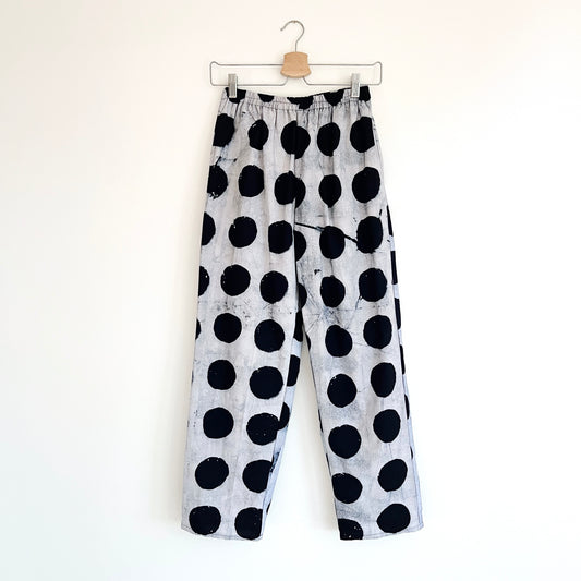 Full front view of Lazy Pants in Indigo Dots Batiki cotton, hanging on a clip hanger against a white wall, showing elastic waistband, bold hand-printed oversized black polka dots on a grey-white ground, and relaxed wide-leg silhouette

