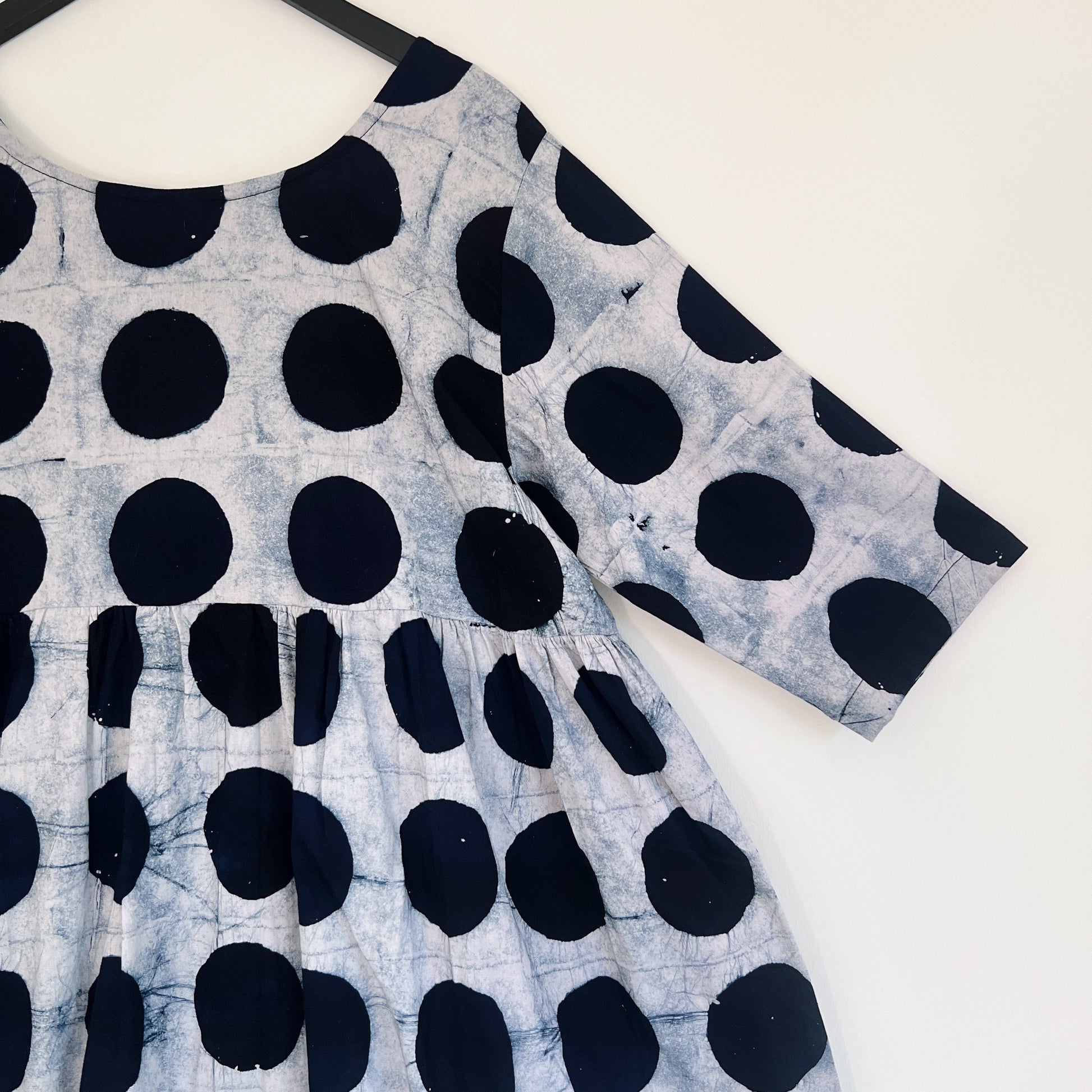 Close-up of the upper bodice and sleeve of the Sula Dress in Black Indigo Dots, highlighting the round neckline, gathered waist seam, elbow-length sleeve, and bold hand-printed black circles on grey-white Batiki cotton with visible hand-dye texture


