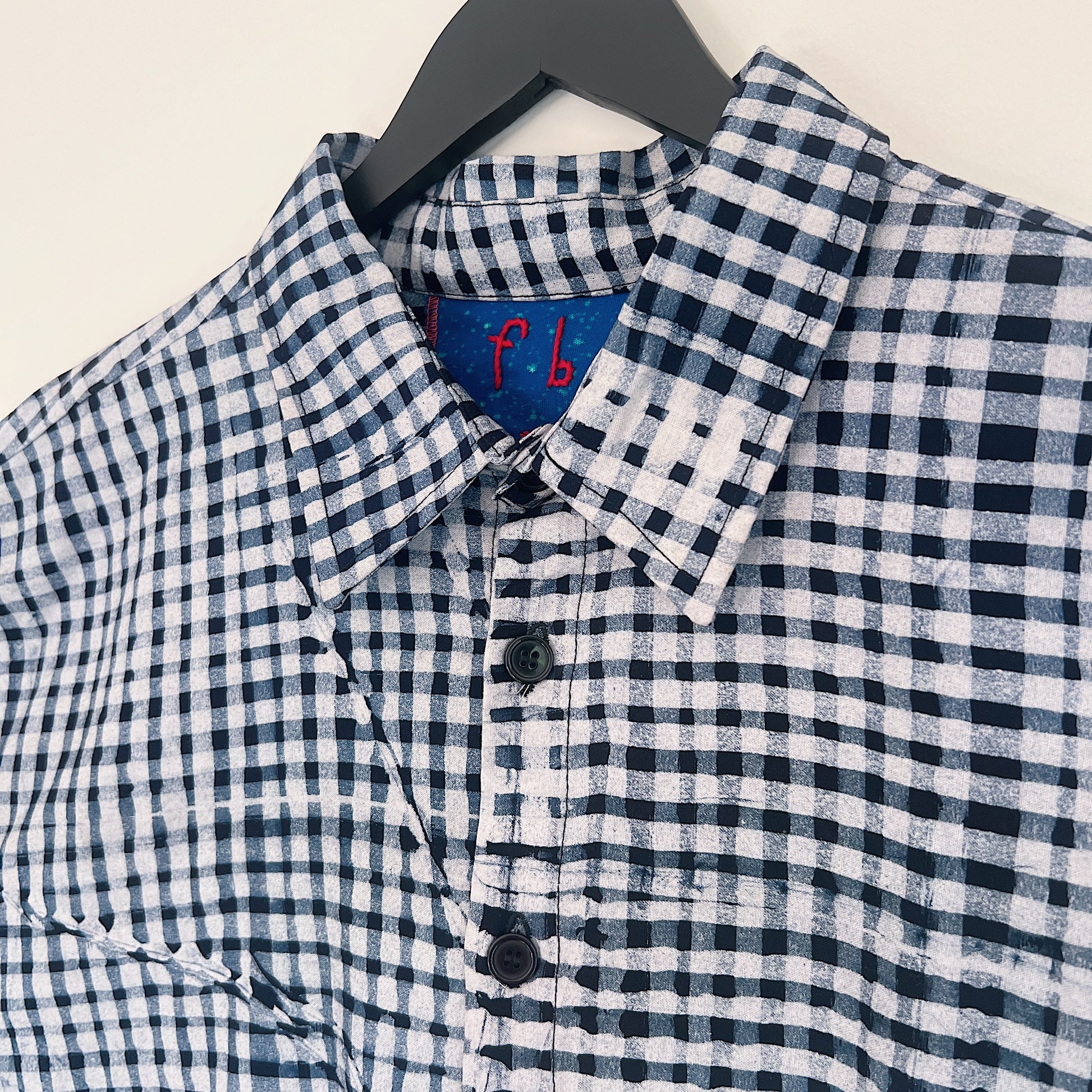 Close-up of the collar and upper chest of the Sudi Shirt in Indigo Grid, showing the point collar with blue Batiki lining and red Fahari Bazaar label, dark buttons, and hand-printed indigo and white gingham grid pattern with natural Batiki dye variation

