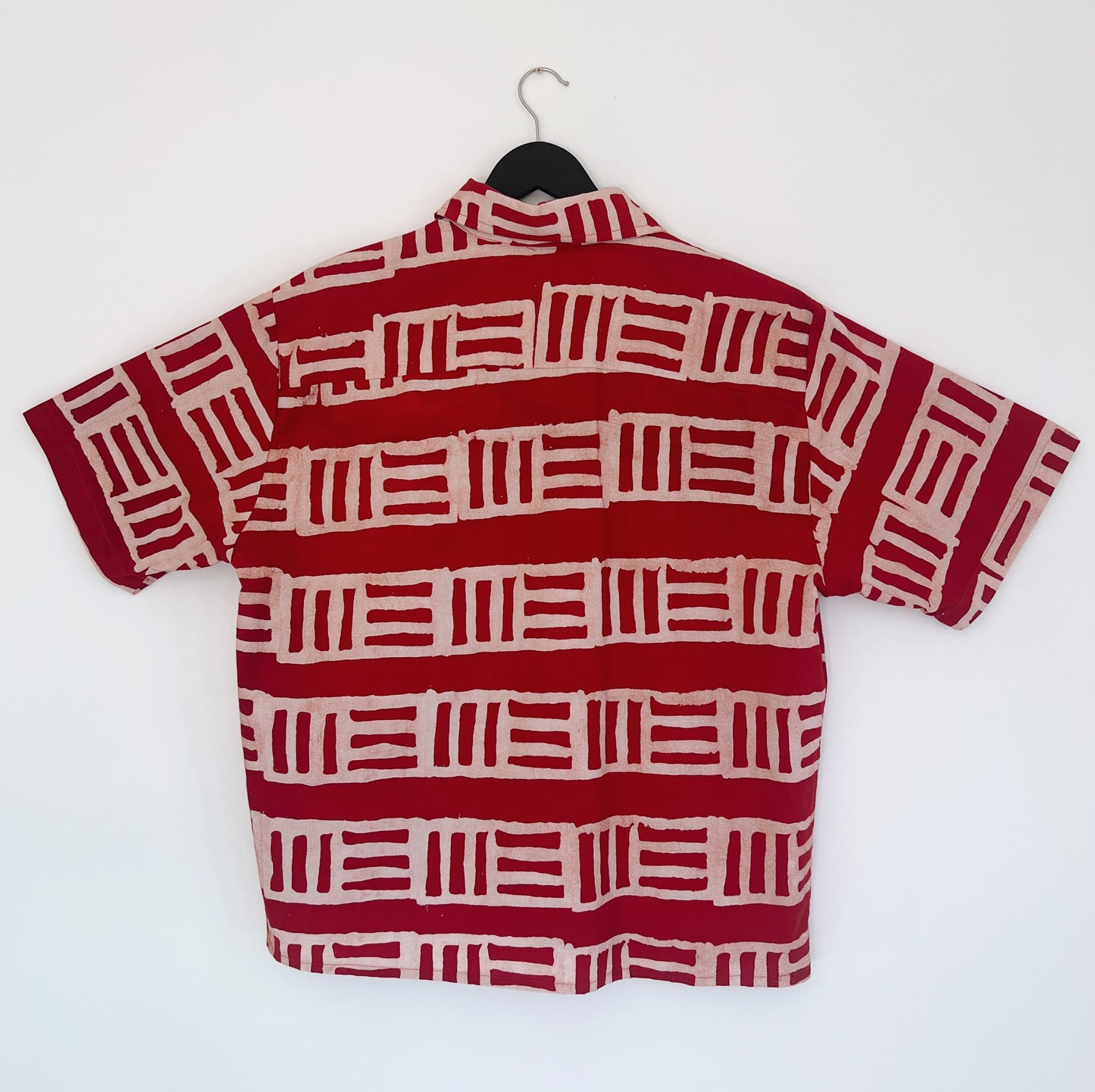 Back view of the Sudi Shirt in Brick, hanging on a black hanger against a white wall, showing the relaxed boxy silhouette, short sleeves, and bold hand-printed brick red and blush geometric block stripe pattern on 100% cotton Batiki fabric

